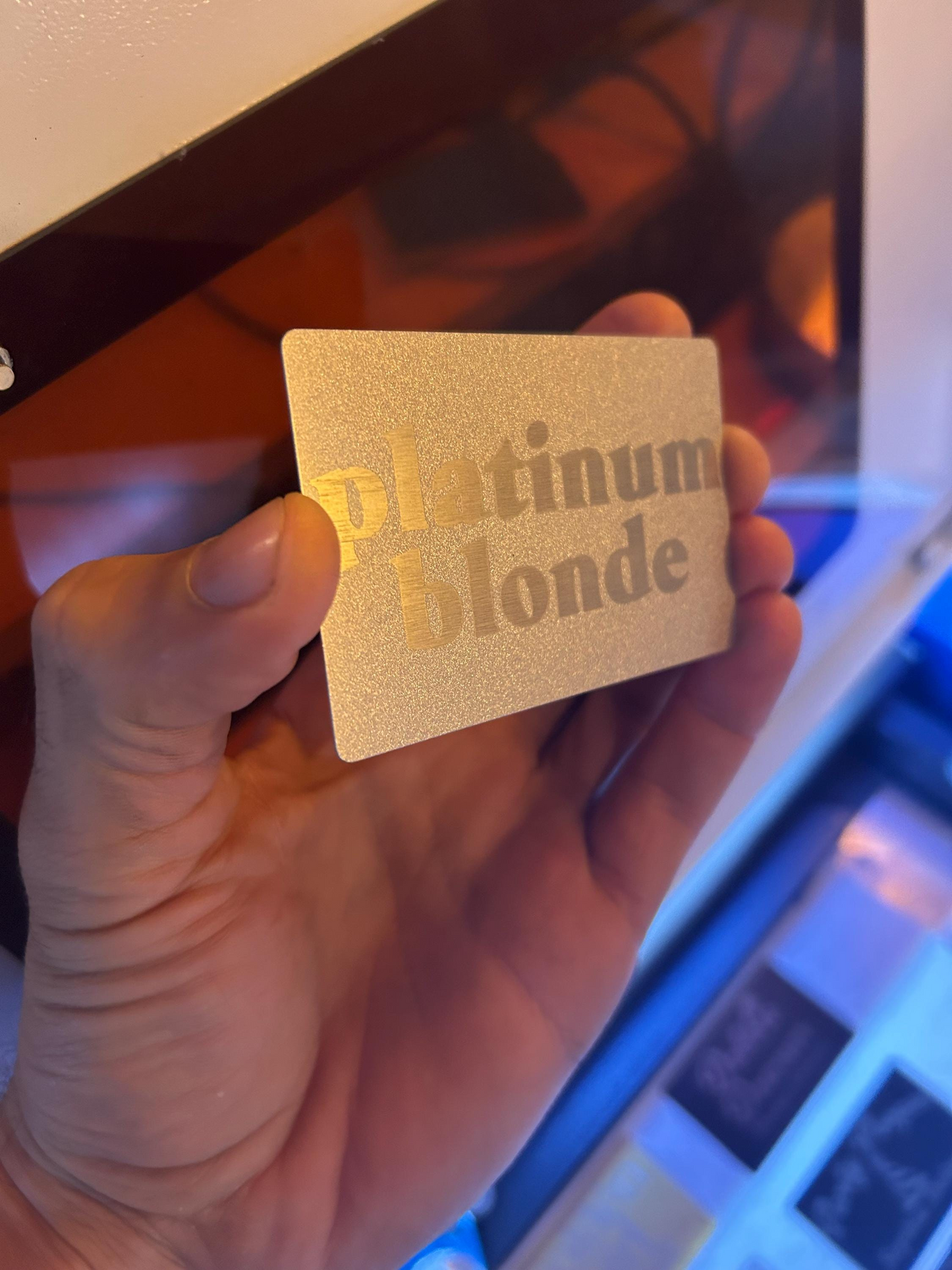 Hand holding a metal silver laser engraved card with 'Platinum Blonde' text in front of a blurred background