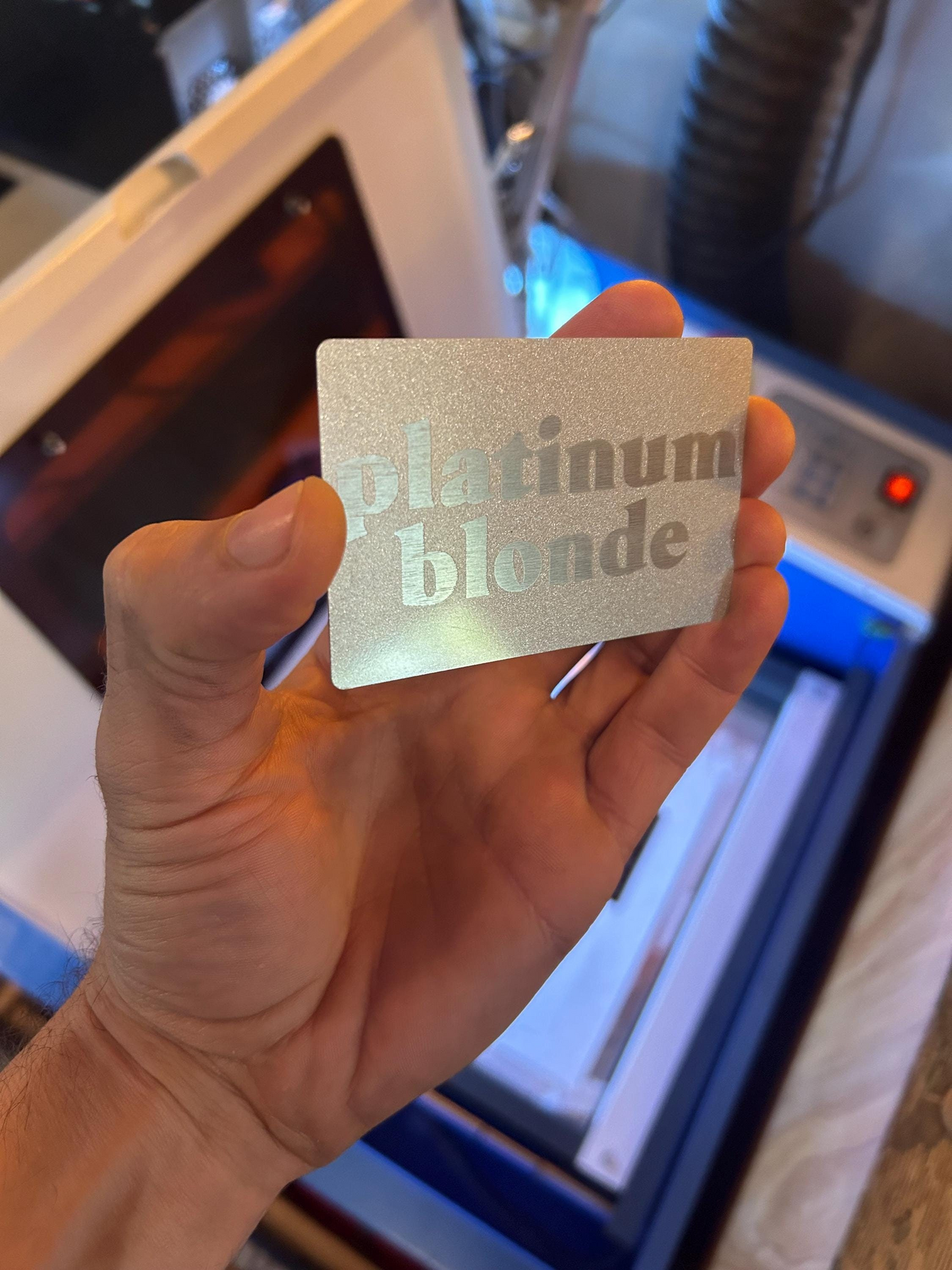 Hand holding a metal keepsake card with 'platinum blonde' laser engraved text in front of a machine.