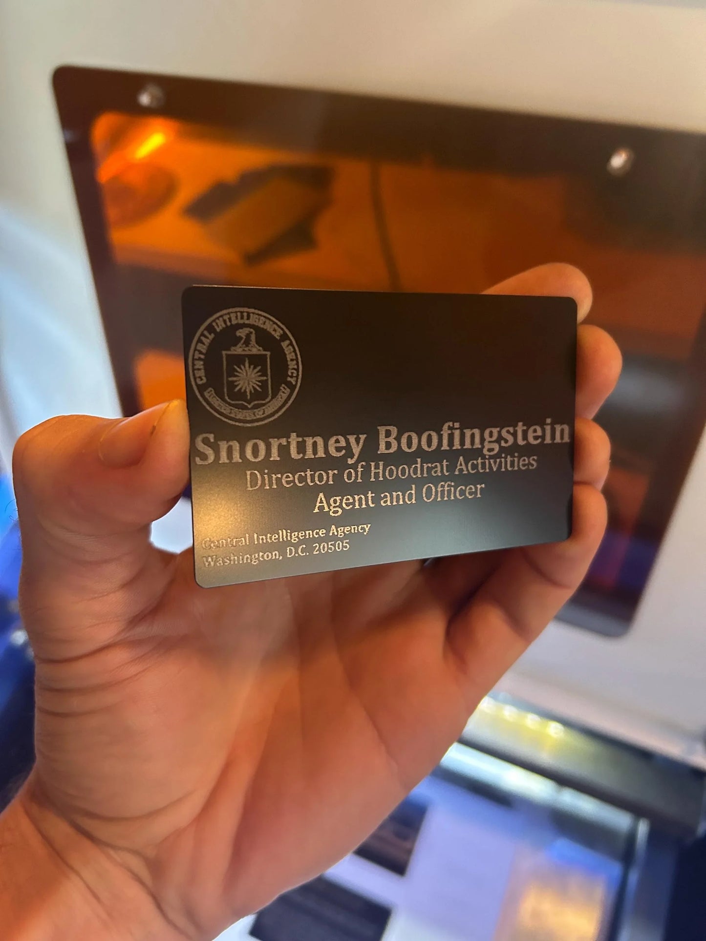 Hand holding a metallic card with text and an emblem, against a blurred indoor background.