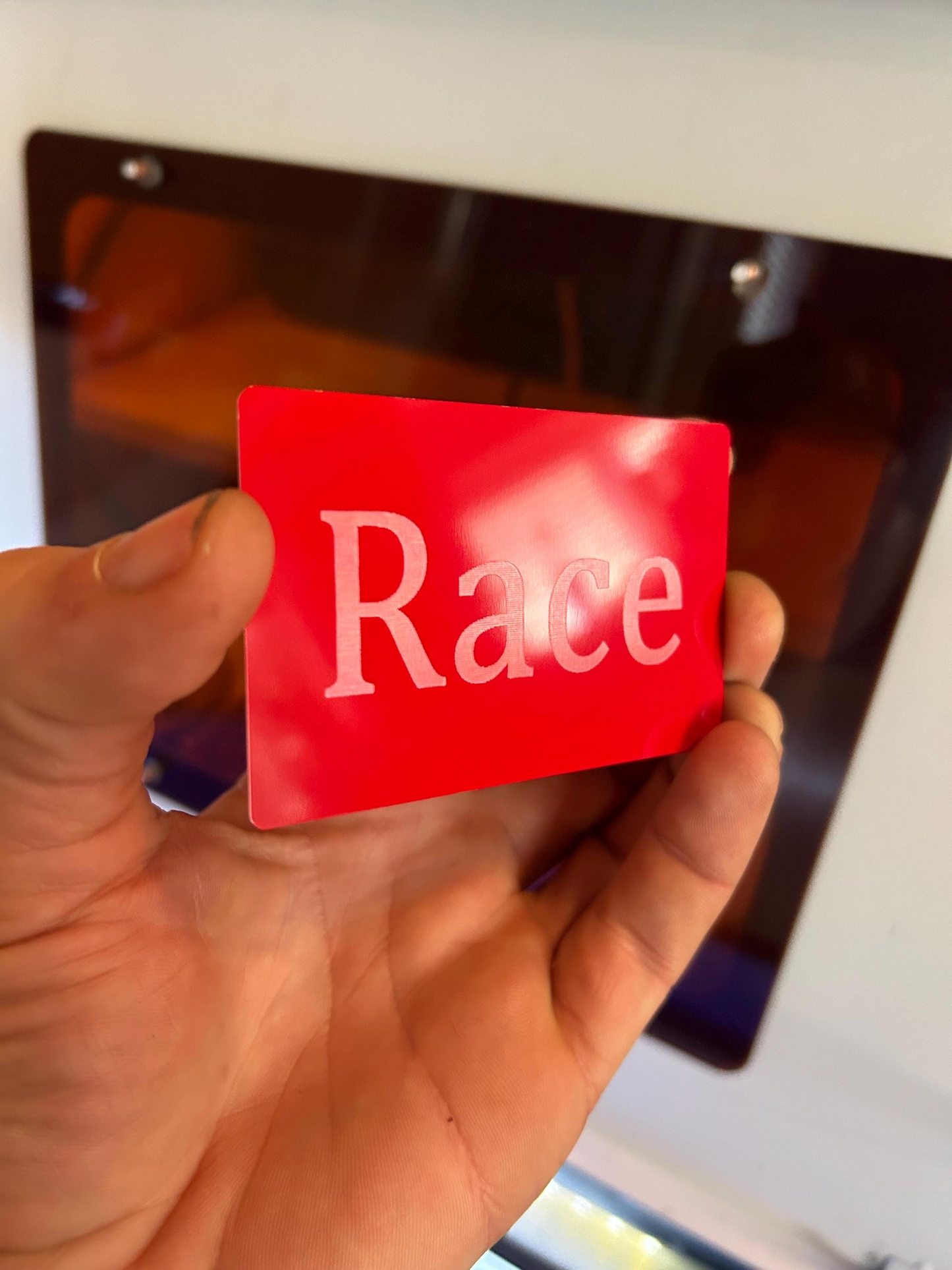 Hand holding a red/silver card with 'Race' laser engraved on it against a blurred background