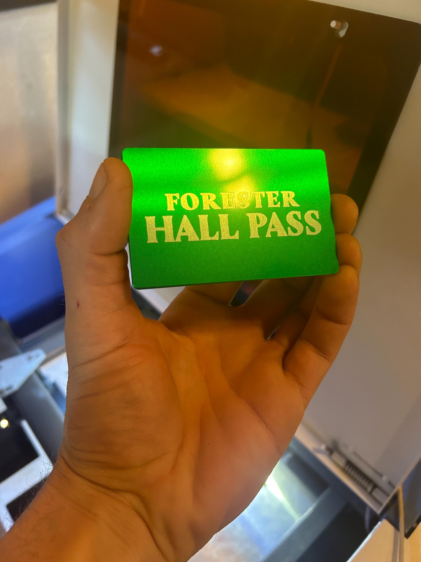 Hand holding a metal green card with customized 'Forever Hall Pass' text in front of a laser machine