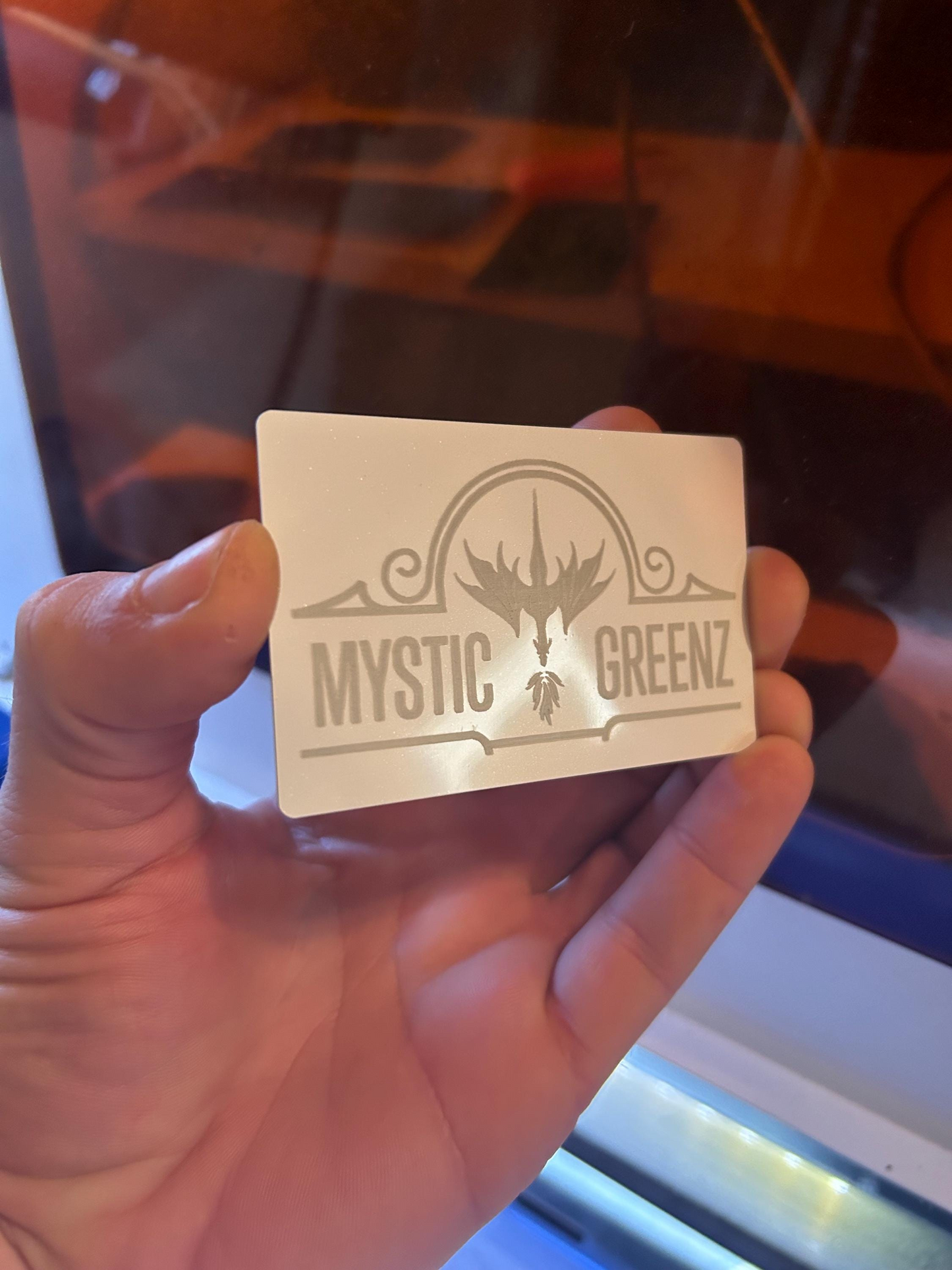 Hand holding a white and silver metal business card, with business logo and dragon imagery in front of a laser cutting machine