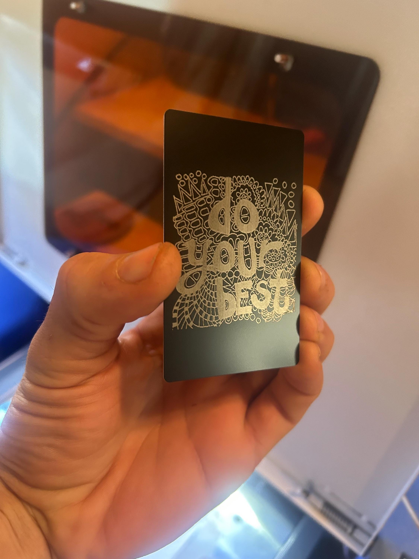 Hand holding an anodized aluminum keepsake card with silver "Do Your Best" text against a blurred background