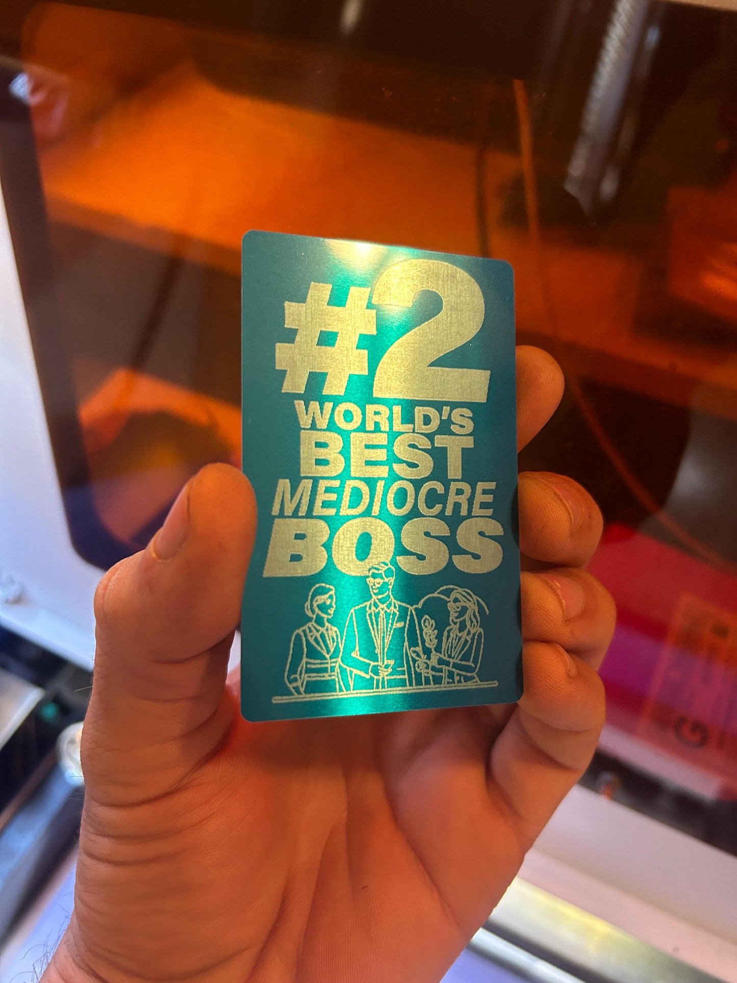 Blue/silver metal card with '#2 World's Best Mediocre Boss' sarcastic funny text held by a hand.