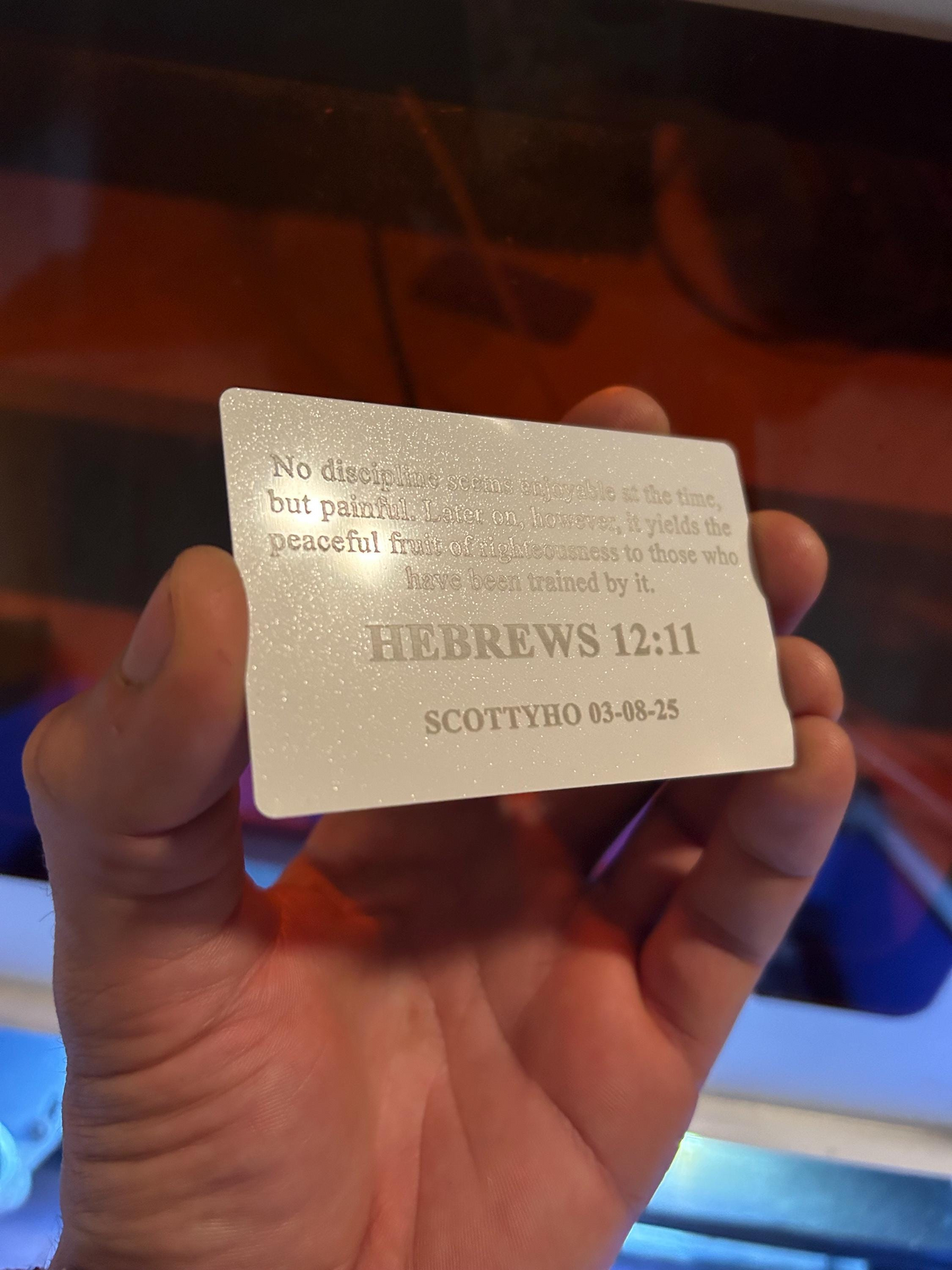 Hand holding a pearl white/silver laser engraved metal bible verse card, with personalized name engraving and Hebrews 12:11 text
