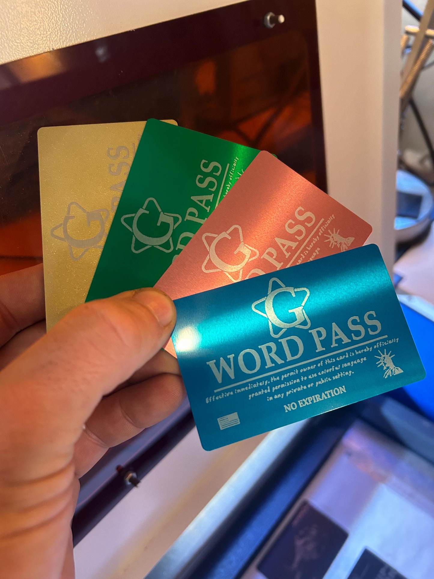 Hand holding a blue, rose gold, green, gold metal novelty gift cards with 'G WORD PASS' laser engraved on each with a production laser engraving machine in the background