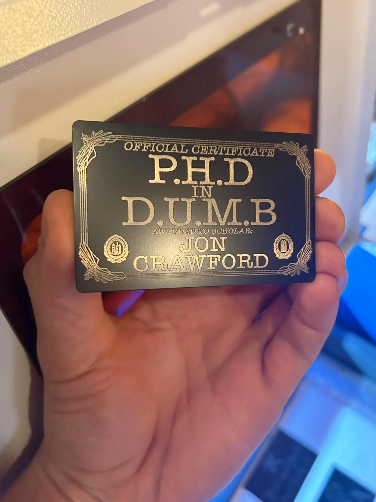 Hand holding a metal keepsake certificate with text 'P.H.D in D.U.M.B' and a personalized name on a blurred background