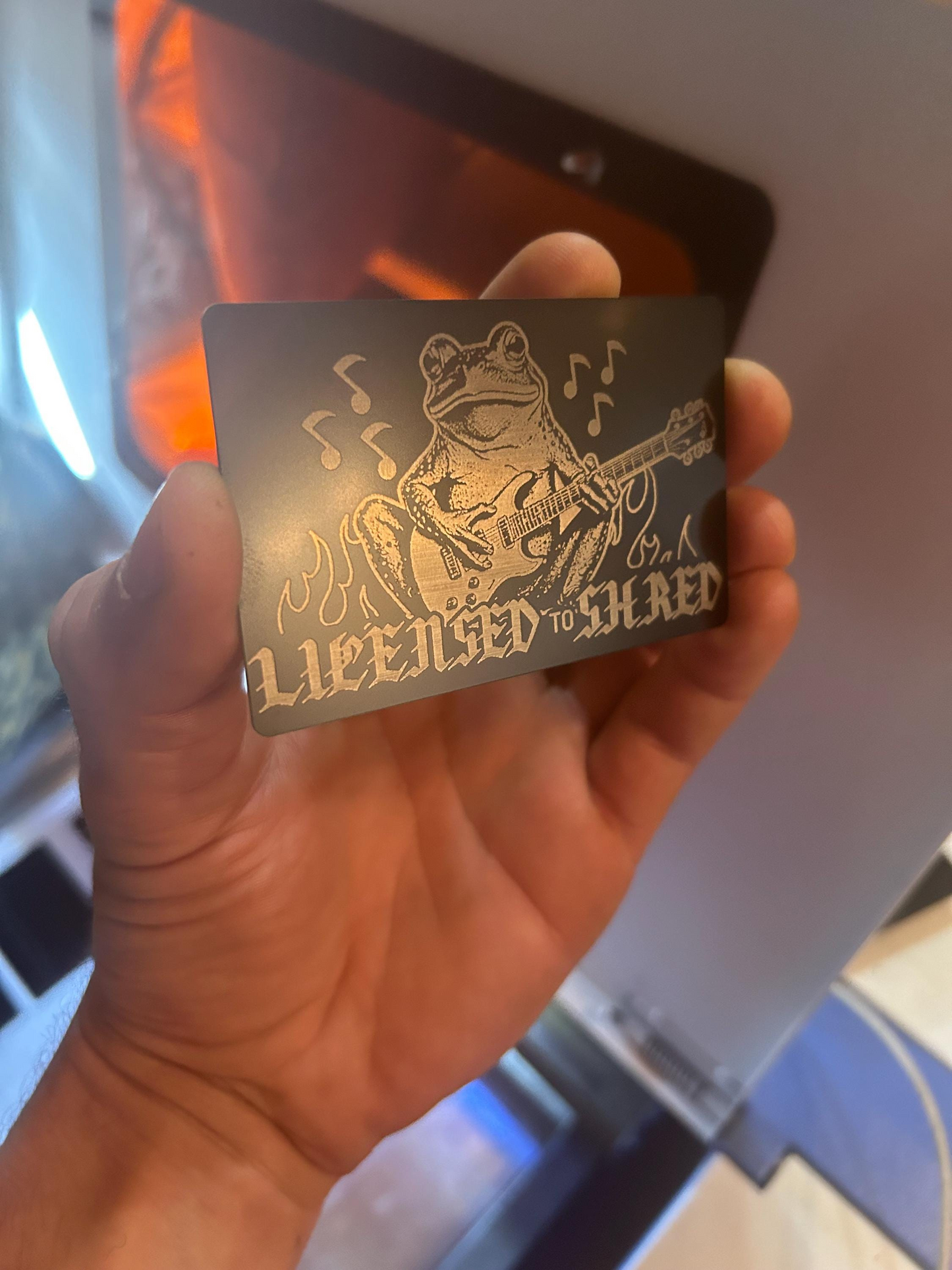 Hand holding an engraved metal placard with a frog graphic and 'Licensed to Shred' text.