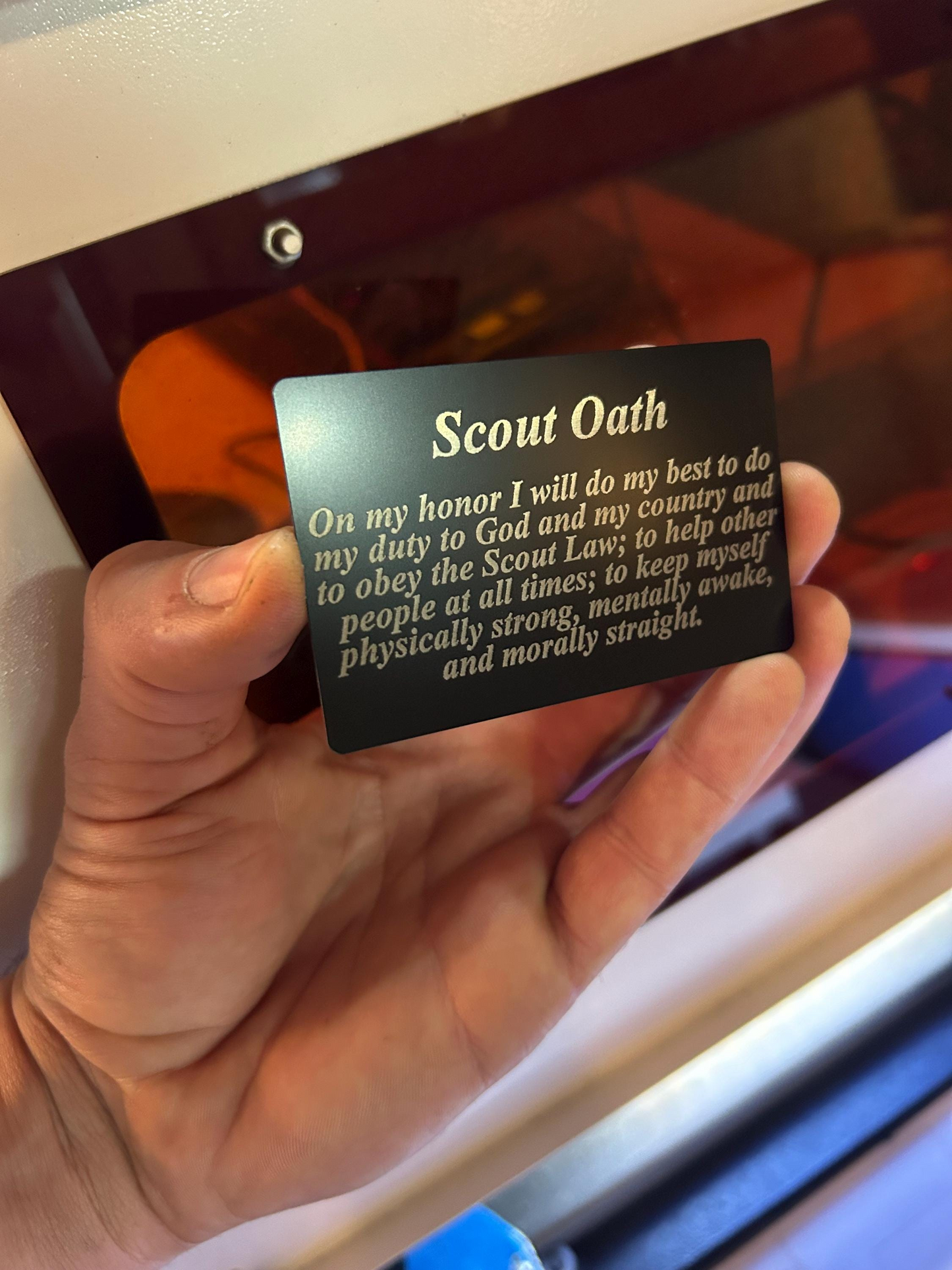 Hand holding a black/silver keepsake placard with 'Scout Oath' text laser engraved for memorization against a blurred background