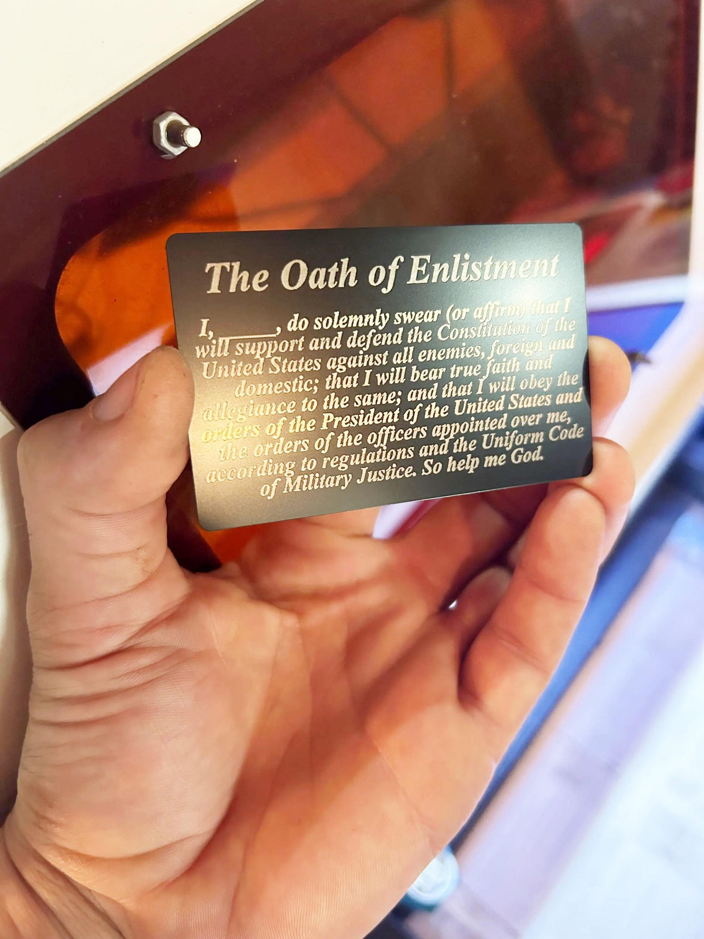 Hand holding a laser engraved metal business card with 'The Oath of Enlistment' text against a blurred background