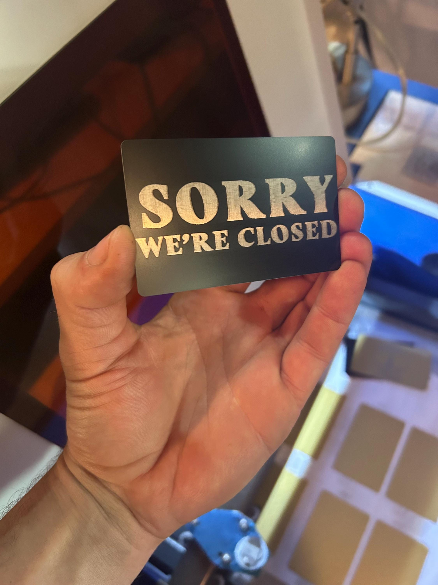 Hand holding a metal black/silver sign that says 'Sorry, We're Closed' in front of a laser engraving machine