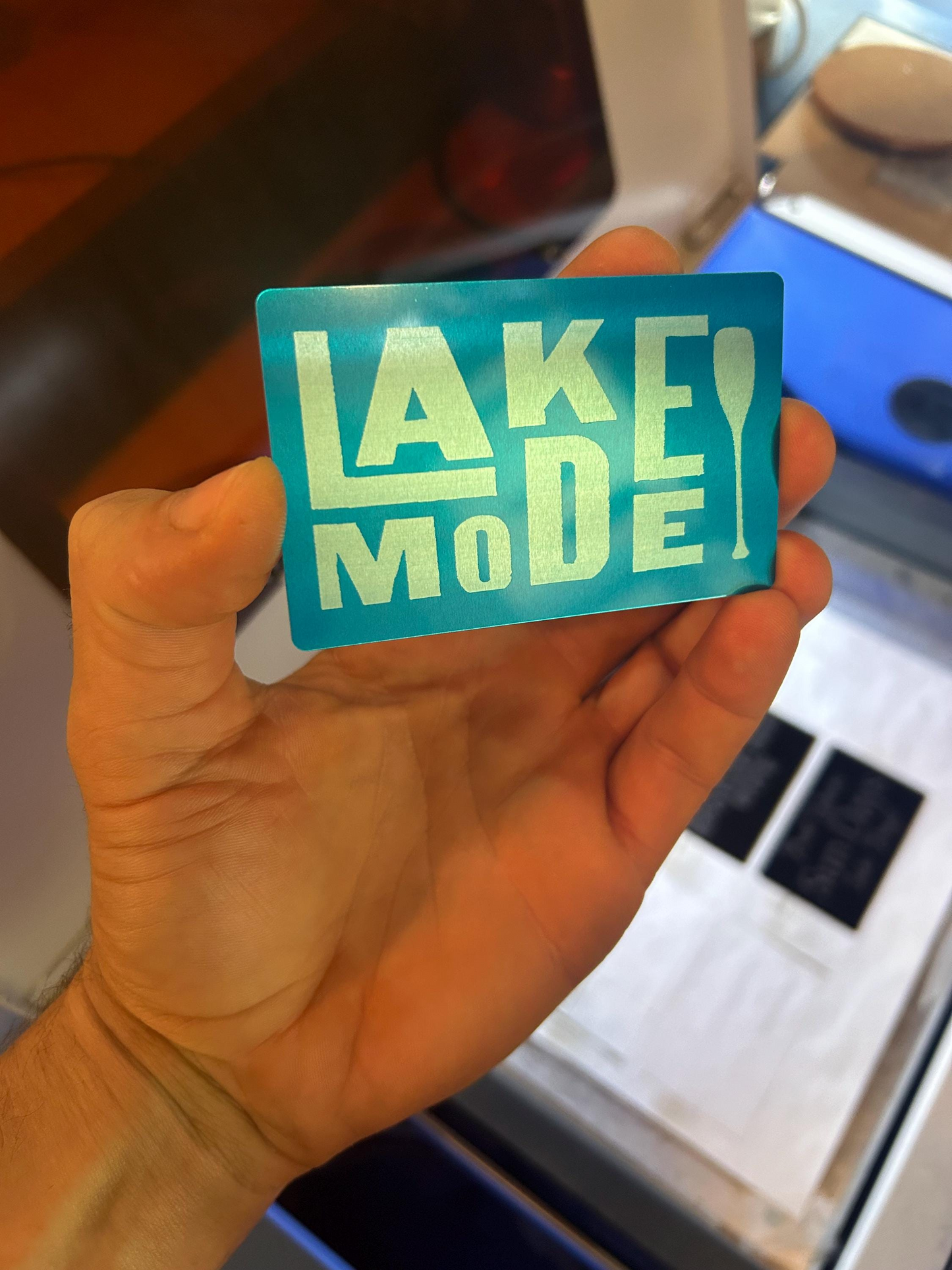 Close-up of hand displaying a laser engraved anodized aluminum lake house card with “Lake Mode” design