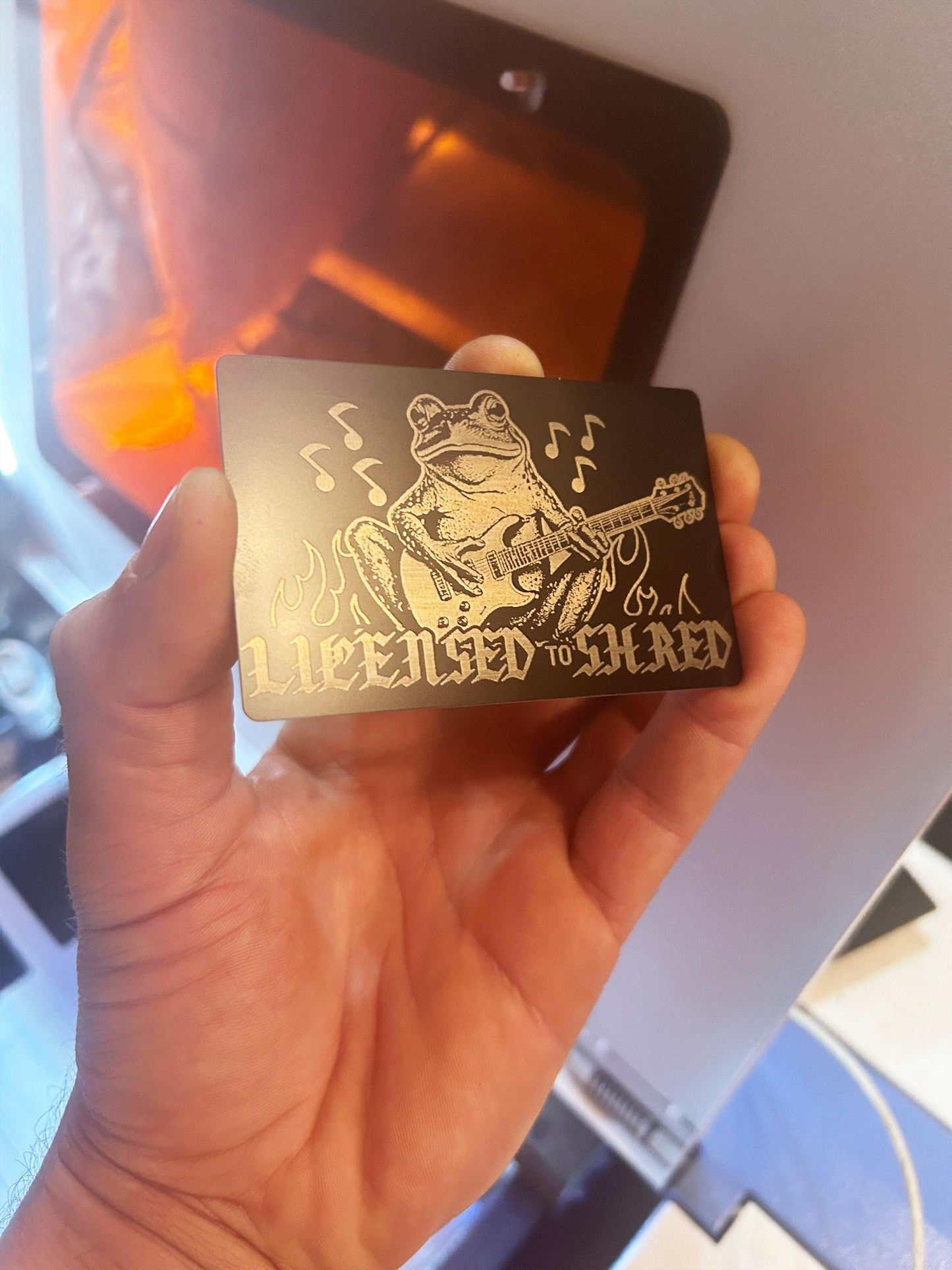 Hand holding a small metal card with a funny frog guitarist design and text, against a blurred background.