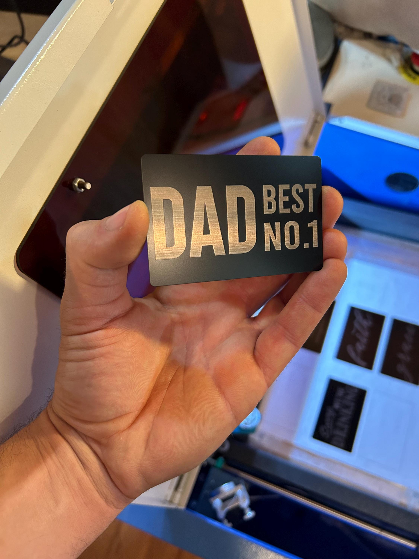 Hand holding a laser engraved wallet card with 'DAD BEST NO.1' text in front of a machine.