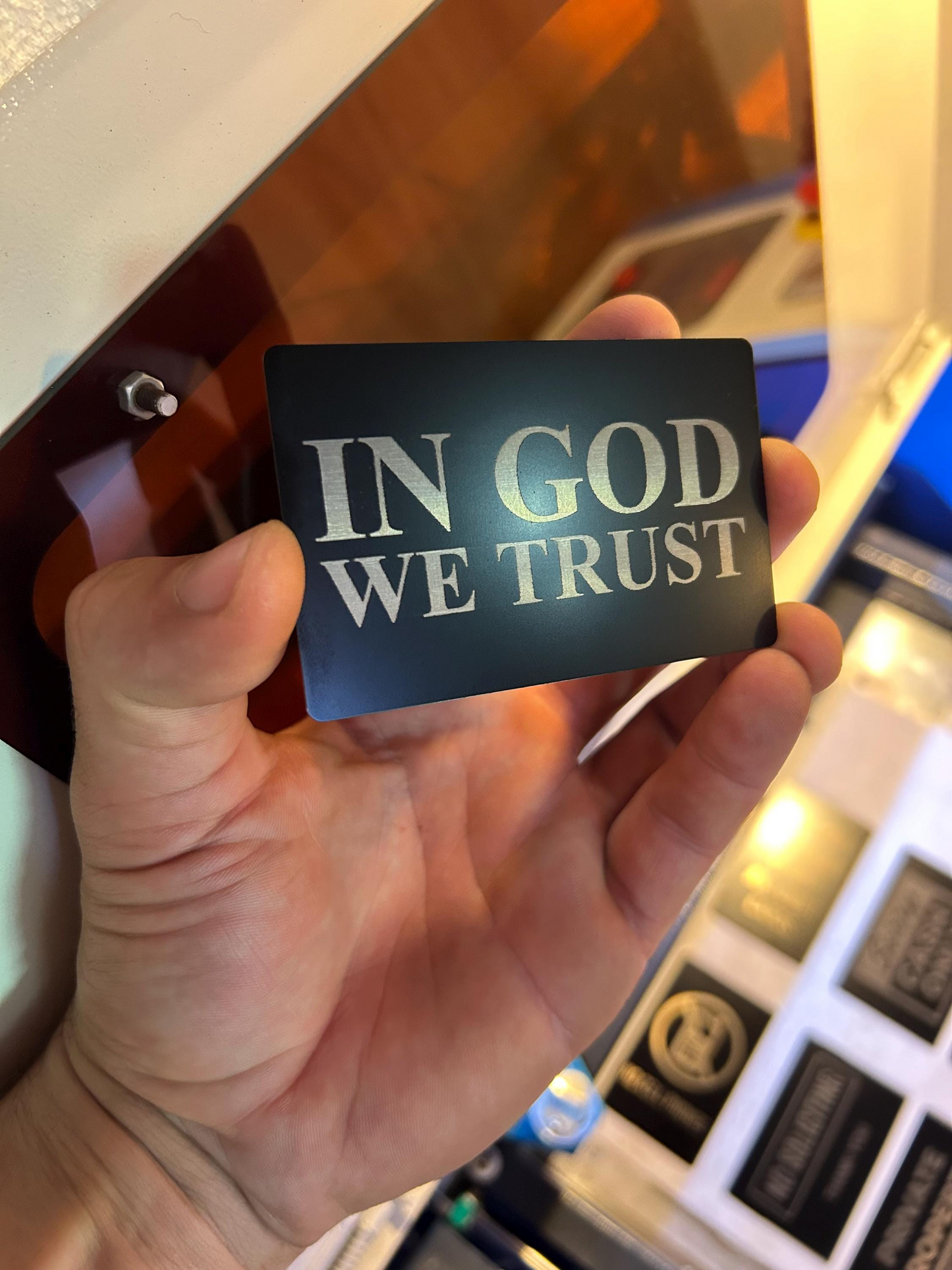 Hand holding a black/silver metal laser engraved bookmark with 'IN GOD WE TRUST' text in front of a blurred background