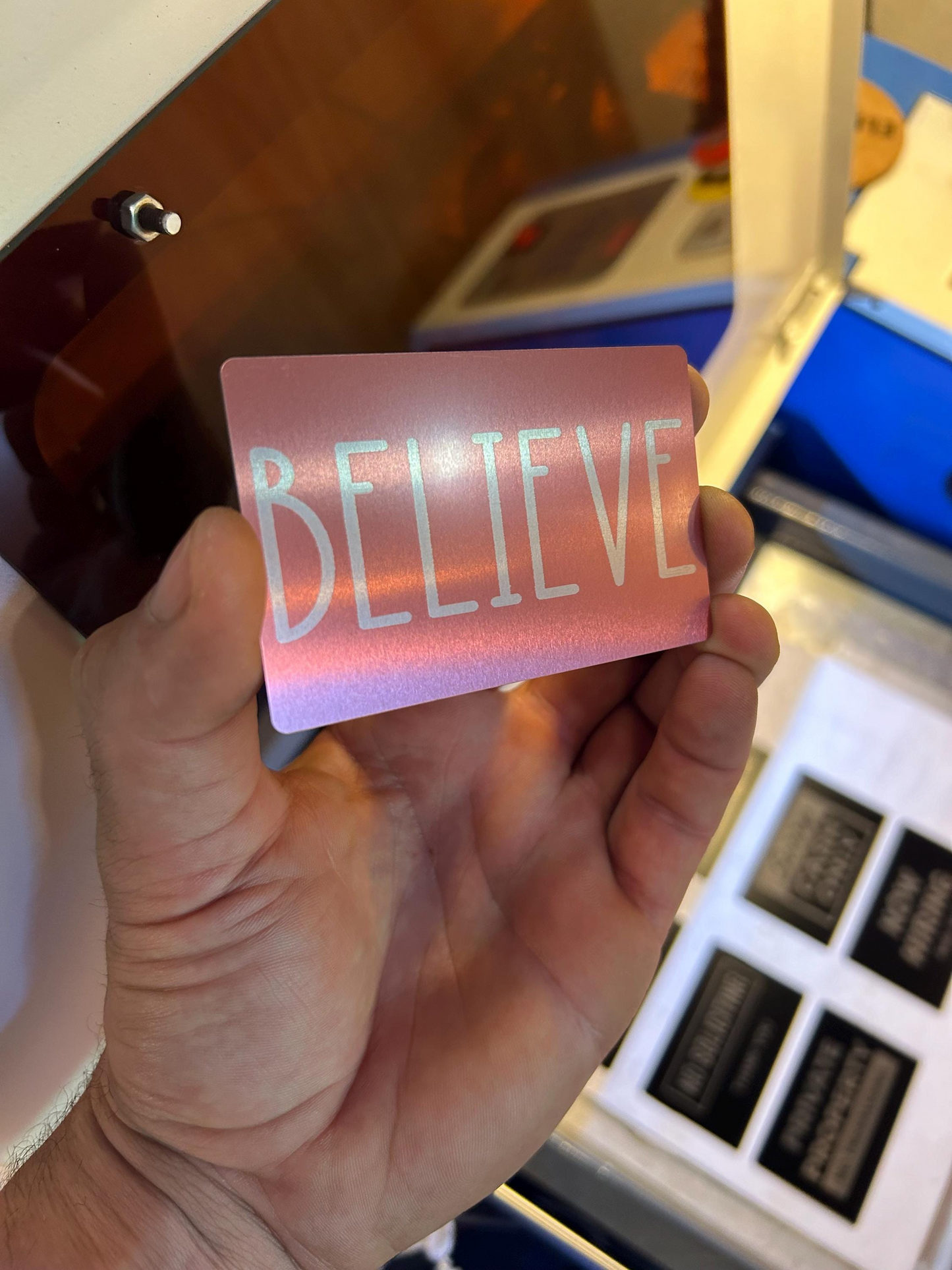 Detailed shot of rose gold anodized metal card engraved with “BELIEVE” text held in hand