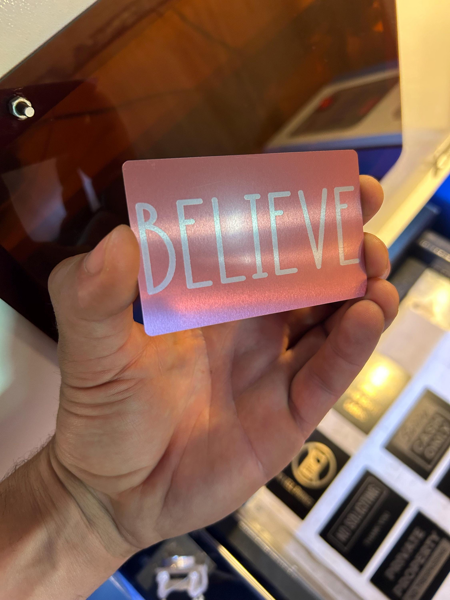 Hand holding a rose gold metal keepsake card with 'BELIEVE' text in front of a blurred background