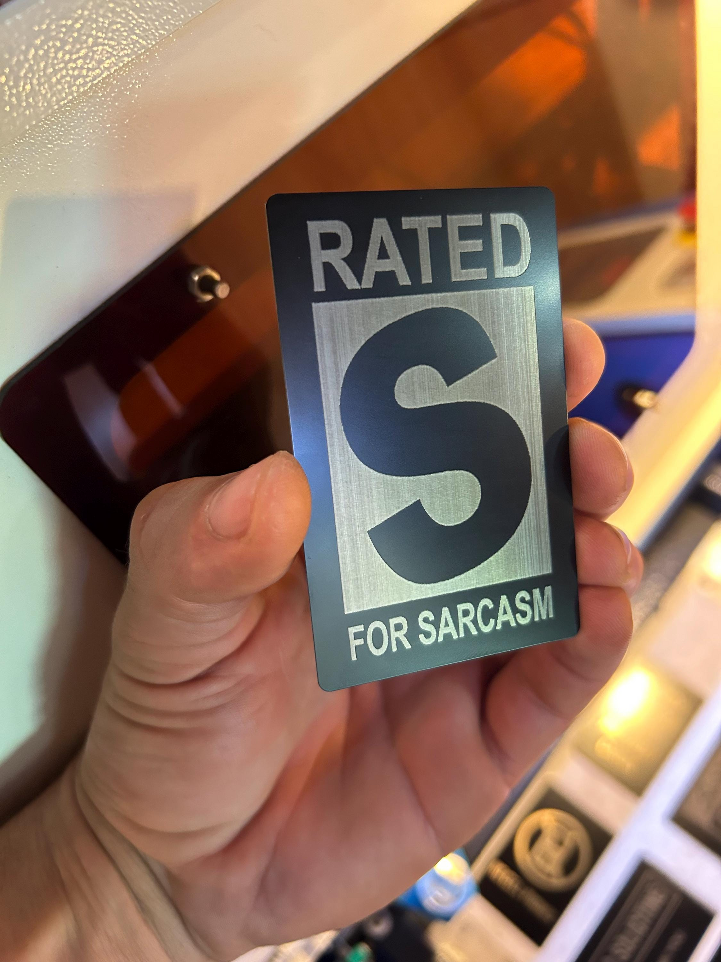 Hand holding a metal keepsake wallet card with 'RATED S FOR SARCASTIC' text against a blurred background
