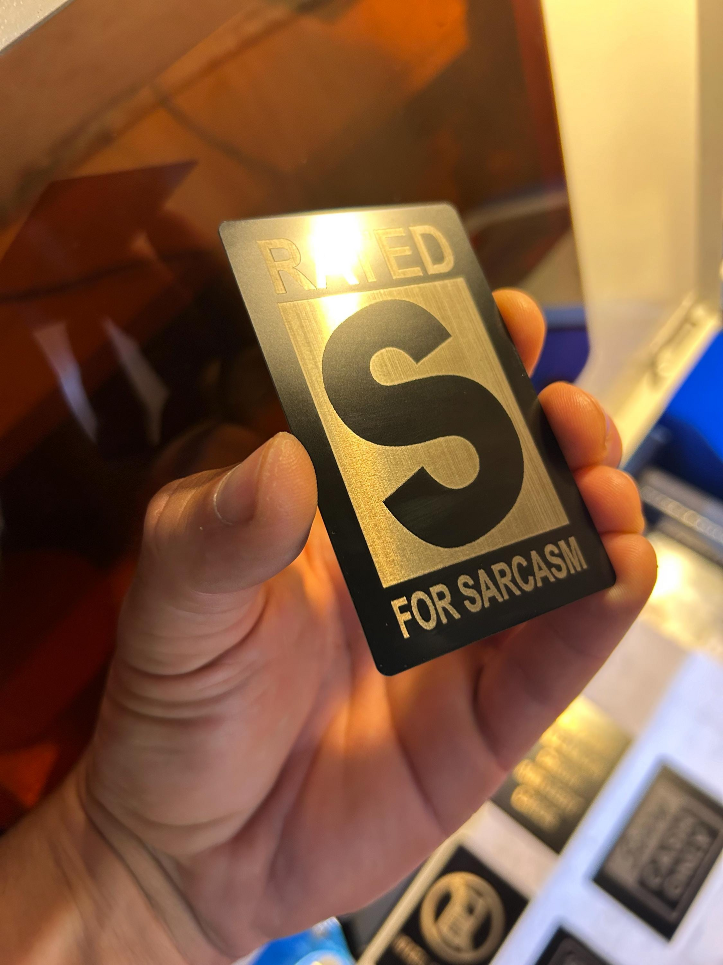 Hand holding a metal gag gift card with 'Rated S for Sarcasm' text laser engraved