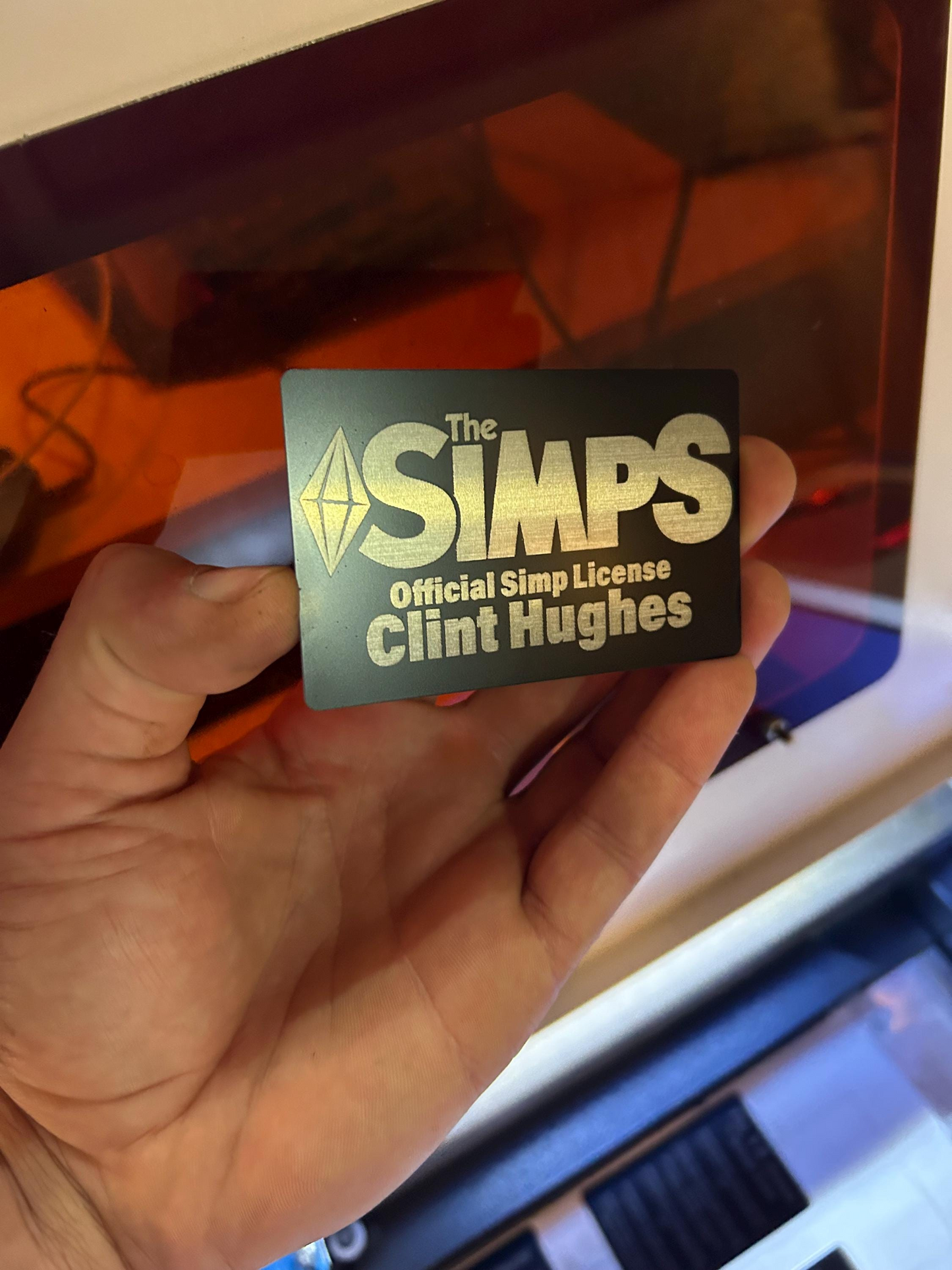 Hand holding a metal 'The Simps' license with text 'Official Simp License and personalized name' against a blurred background.