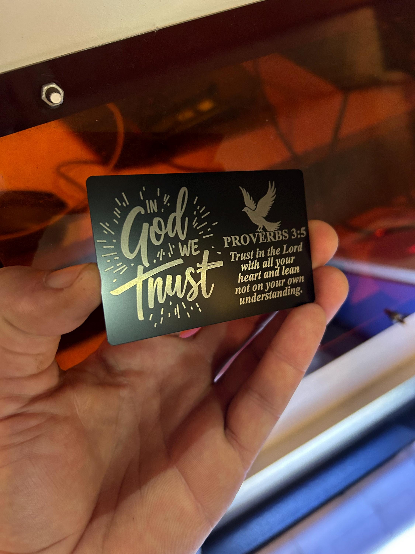 Hand holding a black/silver metal card with 'In God We Trust' and Proverbs 3:5 text.