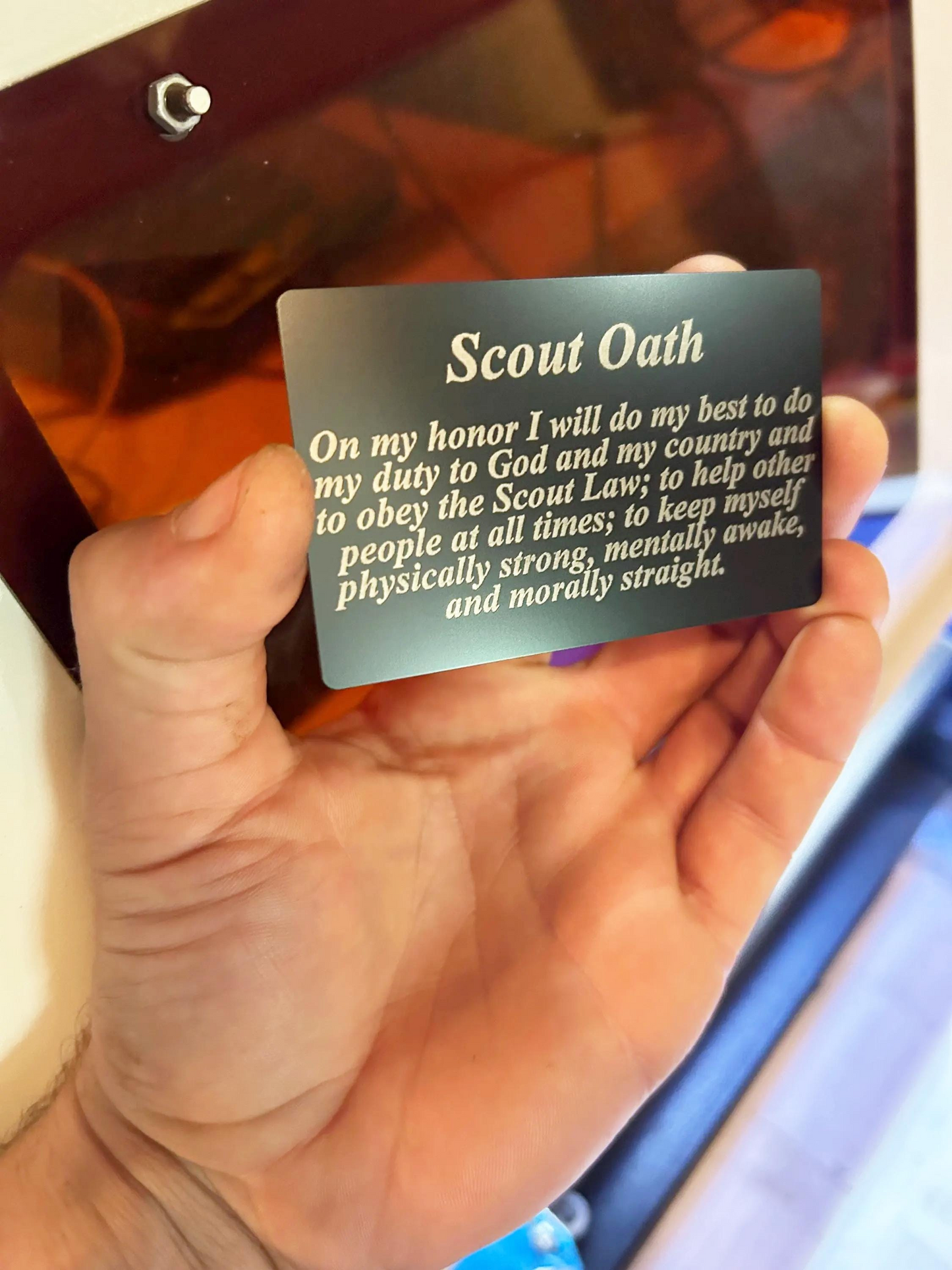 Hand holding a metal black/silver Scout Oath card with laser engraved text on a blurred background