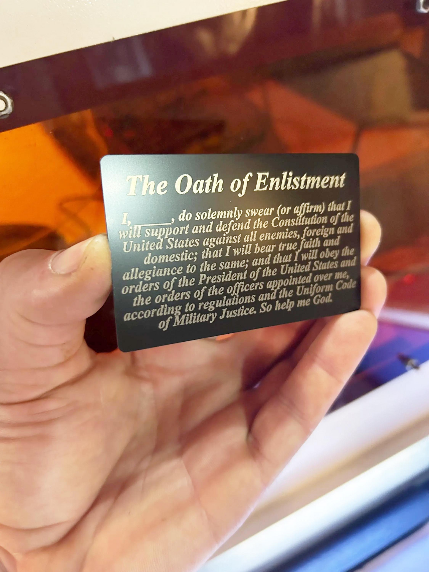 Hand holding a laser engraved metal gift card with 'The Oath of Enlistment' text against a reflective surface.