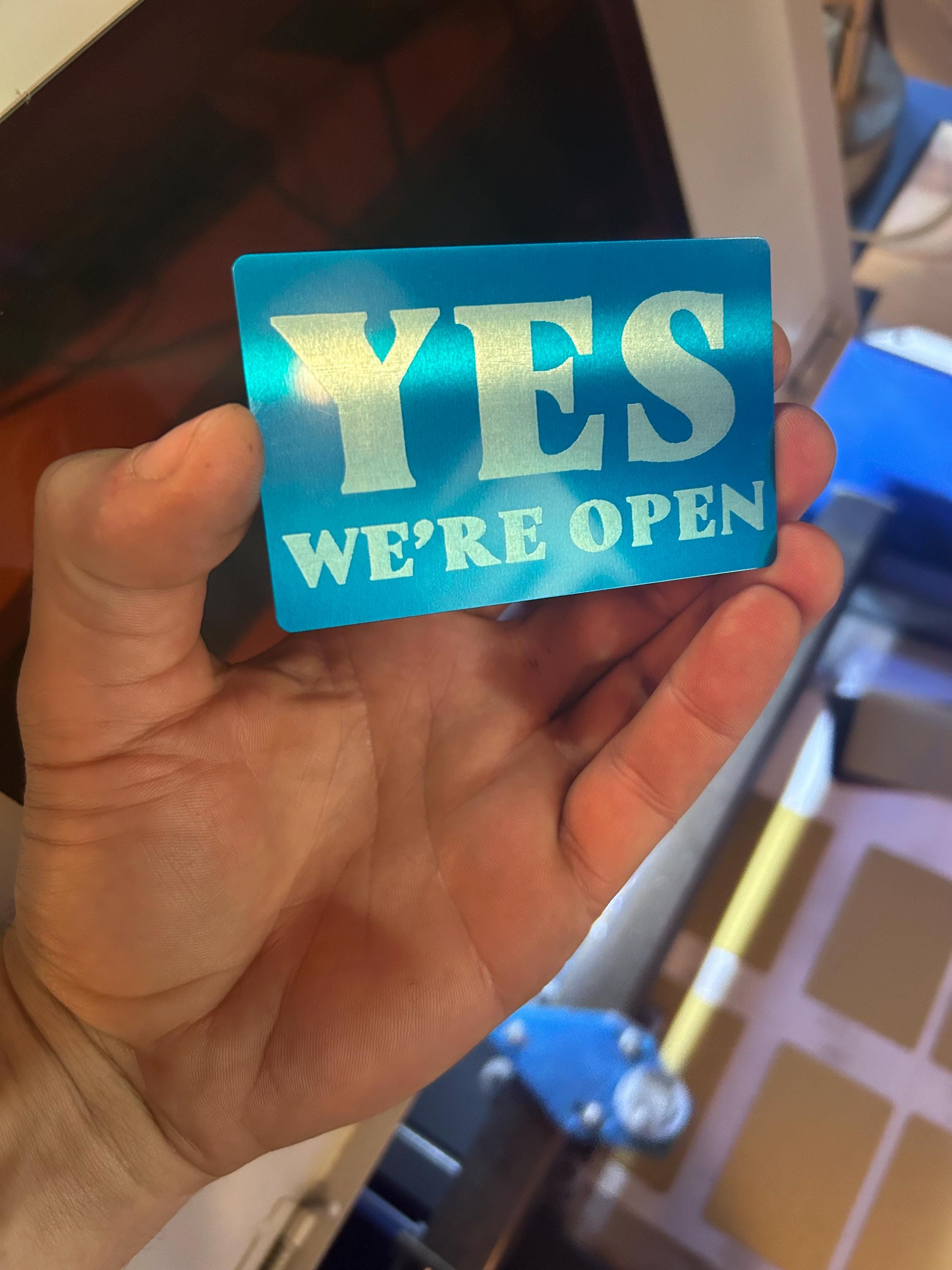 Hand holding a mini metal blue sign with 'YES WE'RE OPEN' text