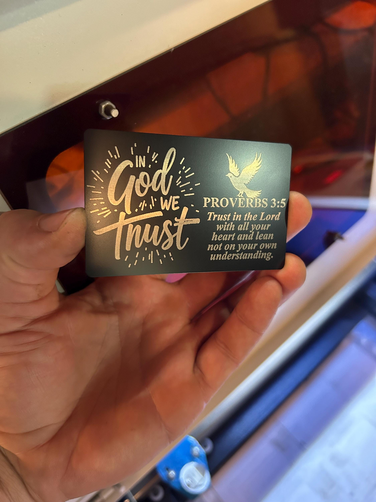 Hand holding a metal laser engraved keepsake card with 'In God We Trust' and Proverbs 3:5 text.