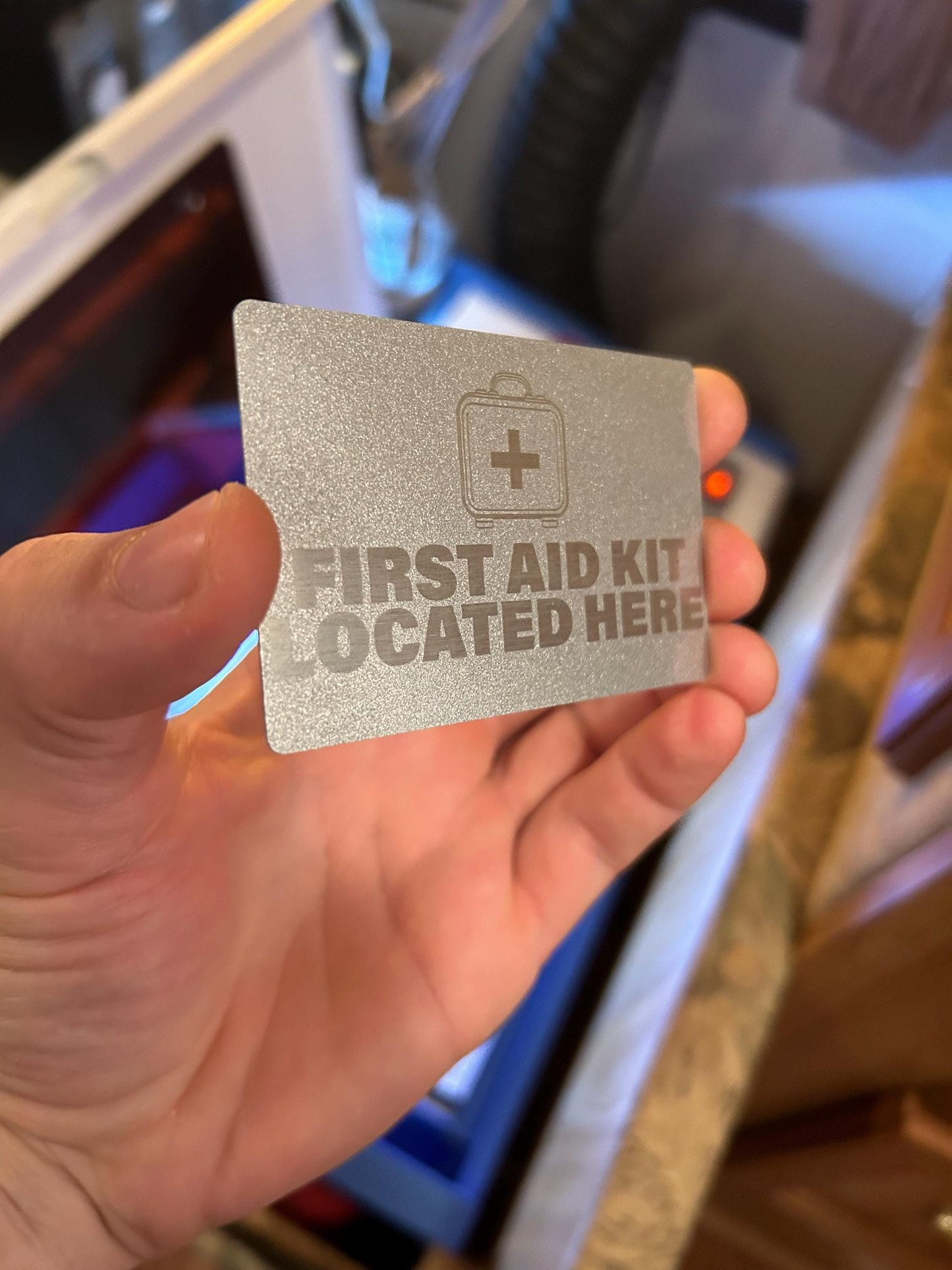 Hand holding a metallic silver 'First Aid Kit Located Here' placard.