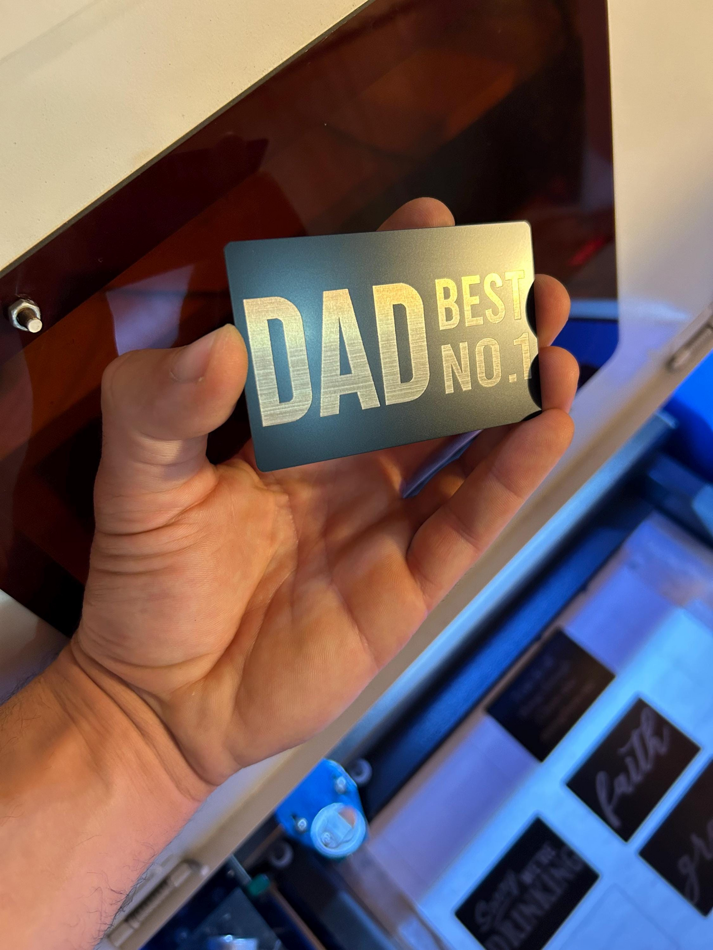 Hand holding a black/silver metal placard with 'DAD BEST NO.1' text in front of a machine.