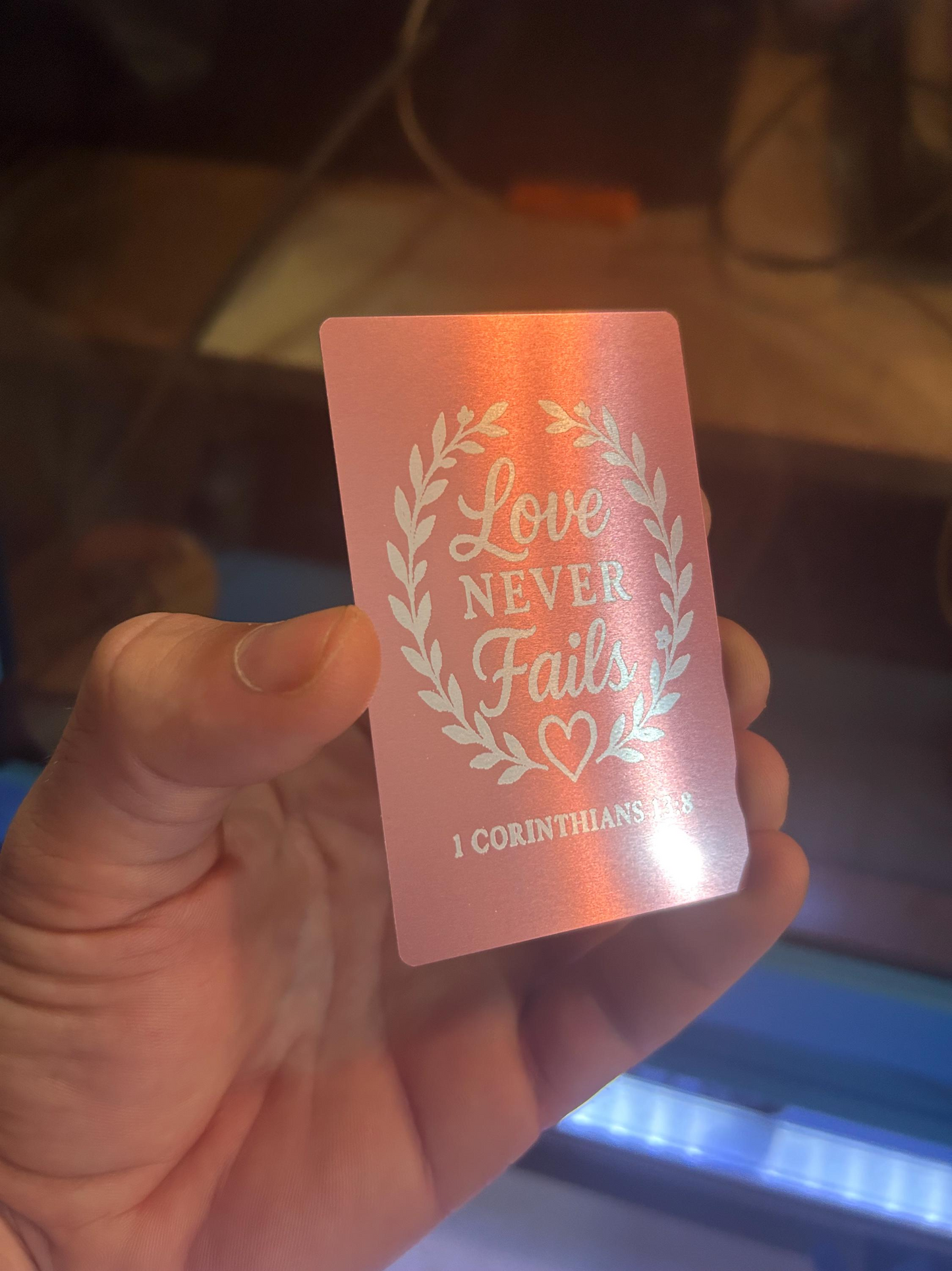 Hand holding laser engraved rose gold inspirational card featuring “Love Never Fails” text with Bible reference below
