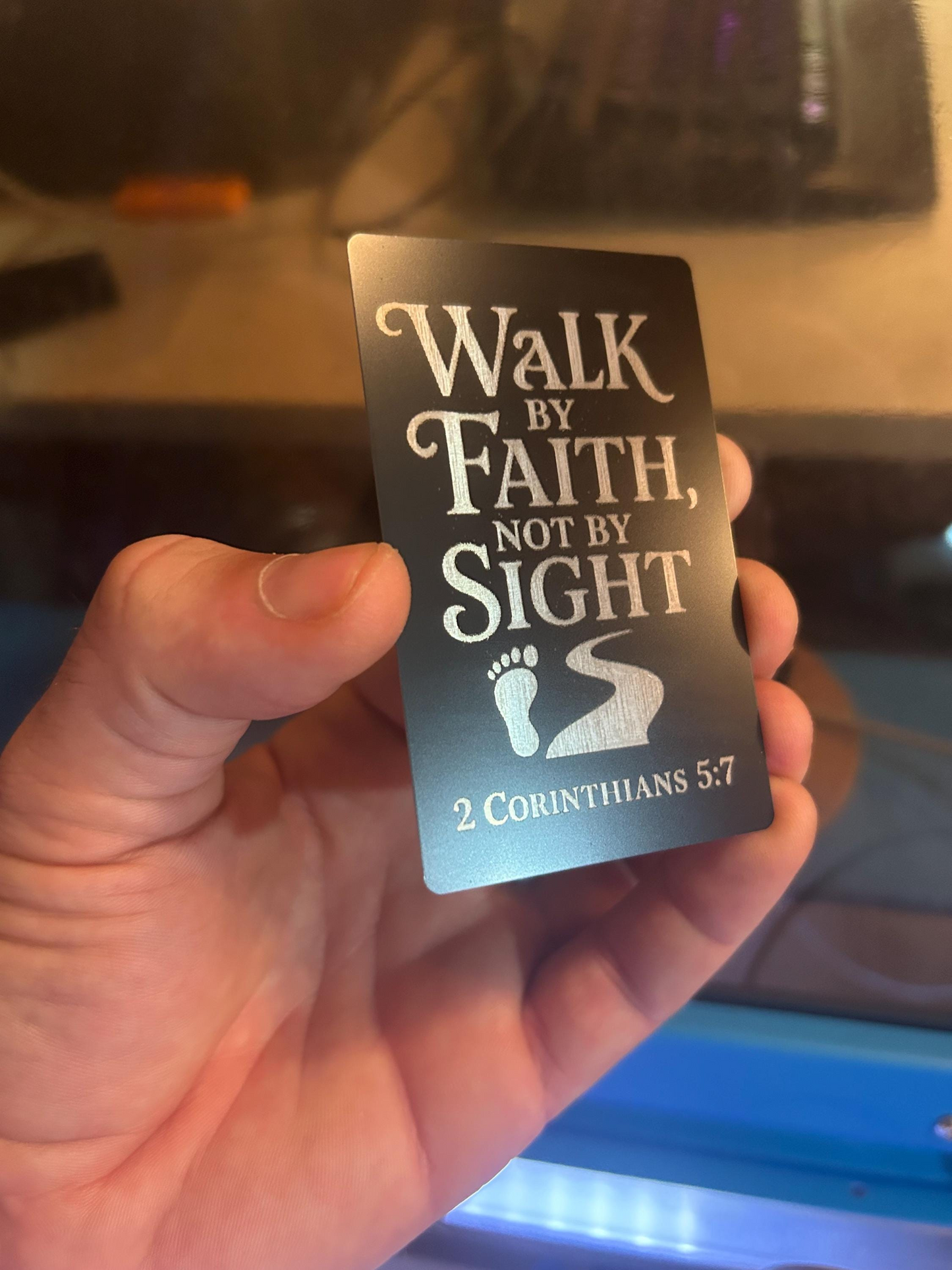 Hand holding a metal bookmark card with 'Walk by Faith, Not by Sight' text and 2 Corinthians 5:7 quote.