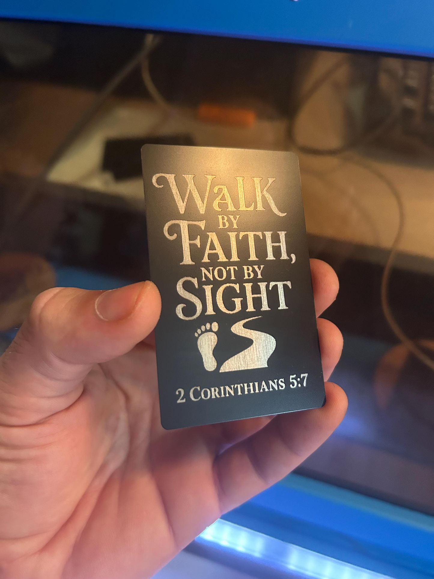 Hand holding a metal keepsake gift card with 'Walk by Faith, Not by Sight' text against a blurred background