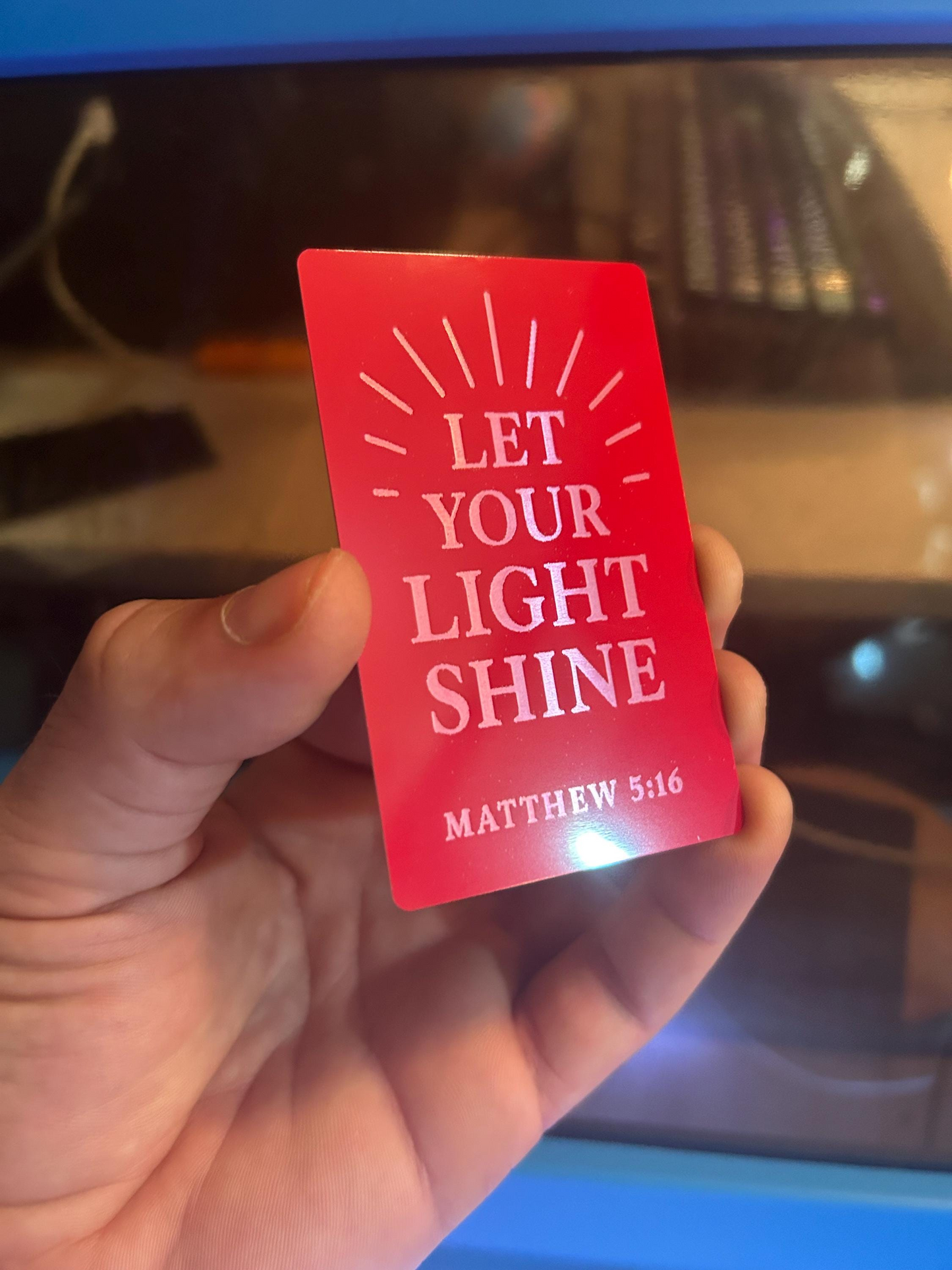 Hand holding a red and silver metal keepsake card engraved with “Let Your Light Shine” and Matthew 5:16 against blurred laser machine background