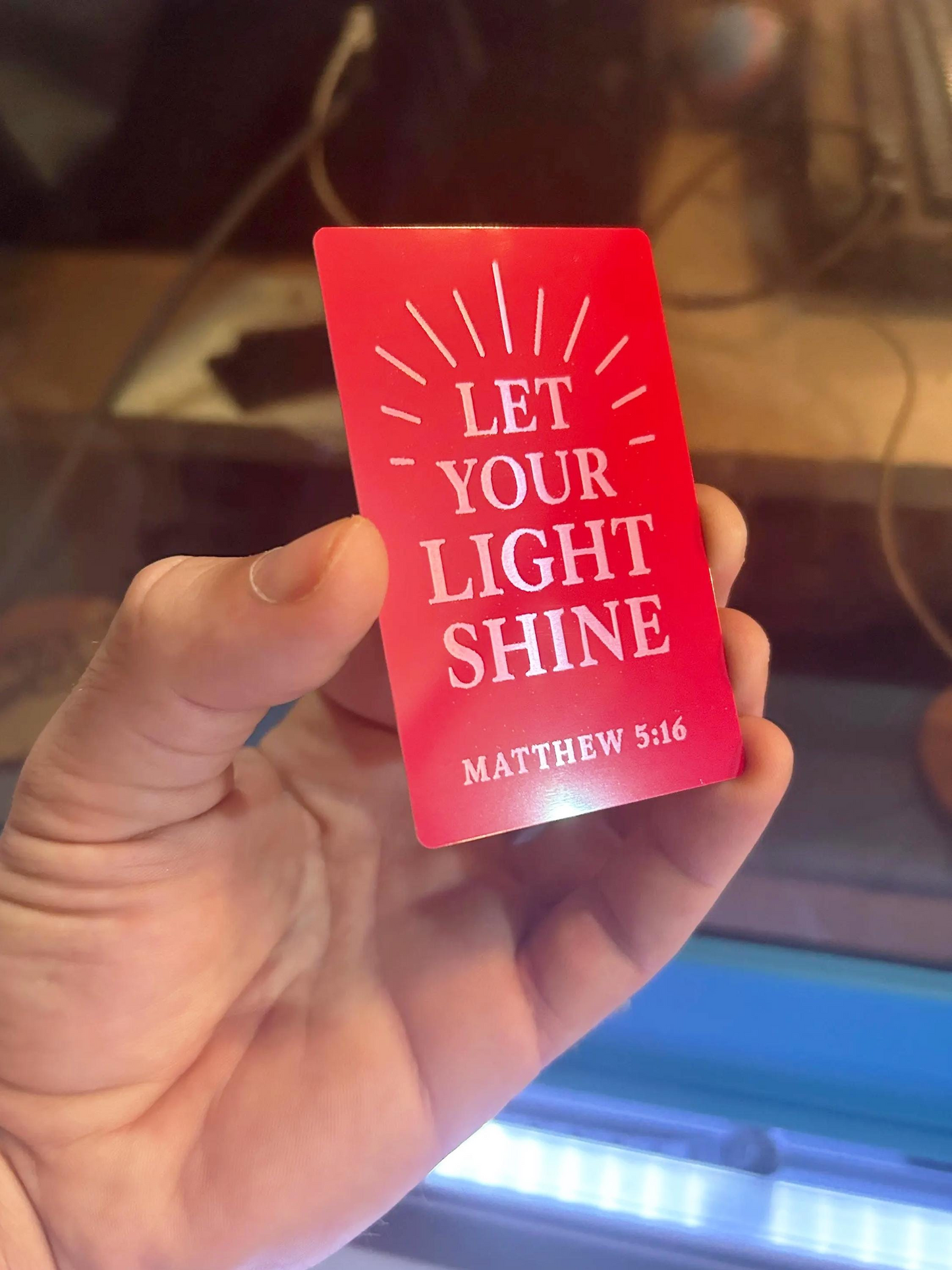 Hand presenting a custom Christian metal keepsake card – “Let Your Light Shine” design with sun rays in red/silver anodized aluminum
