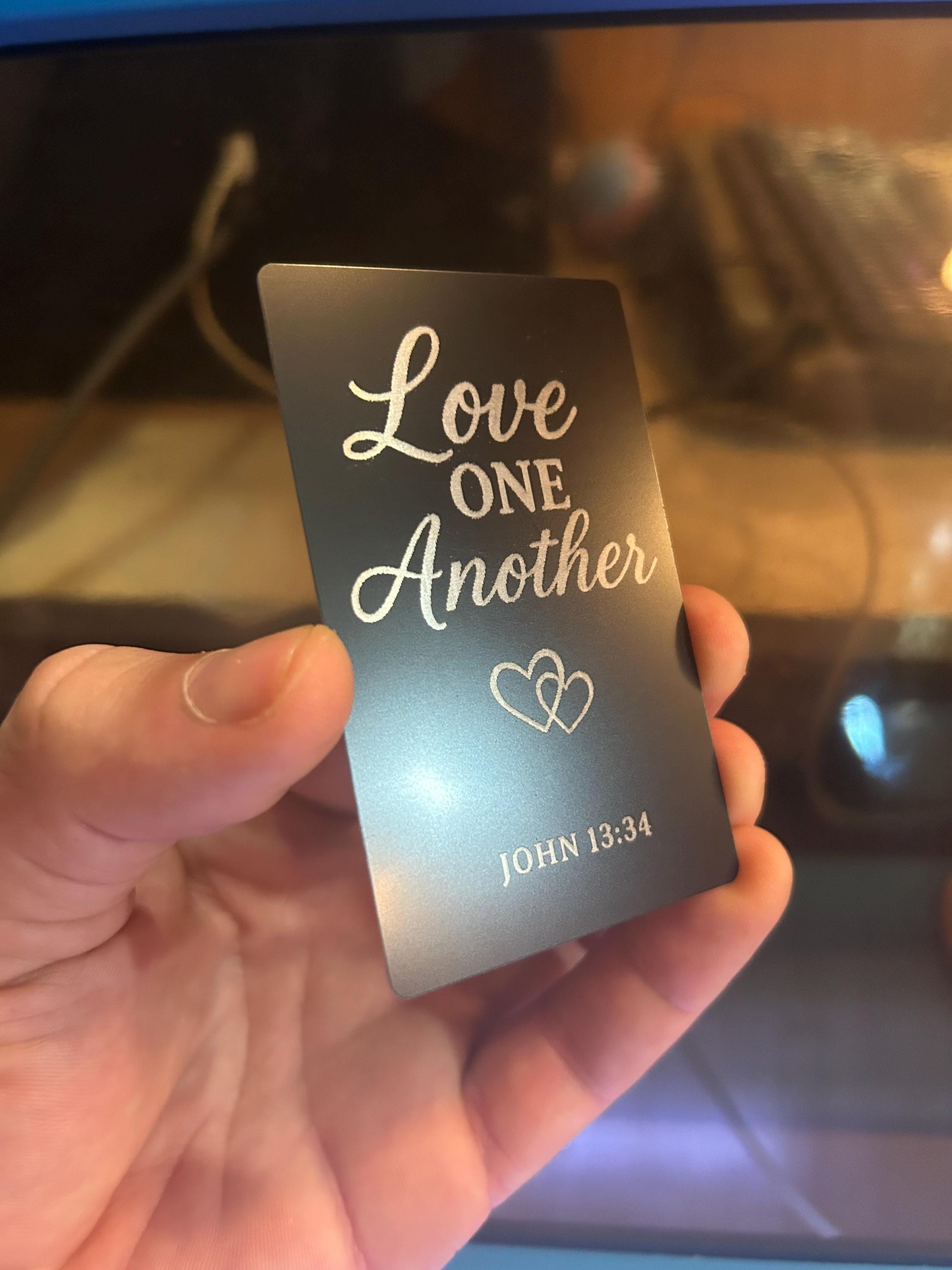 Hand presenting a laser cut black metal religious plaque with engraved “Love One Another” text and John 13:34