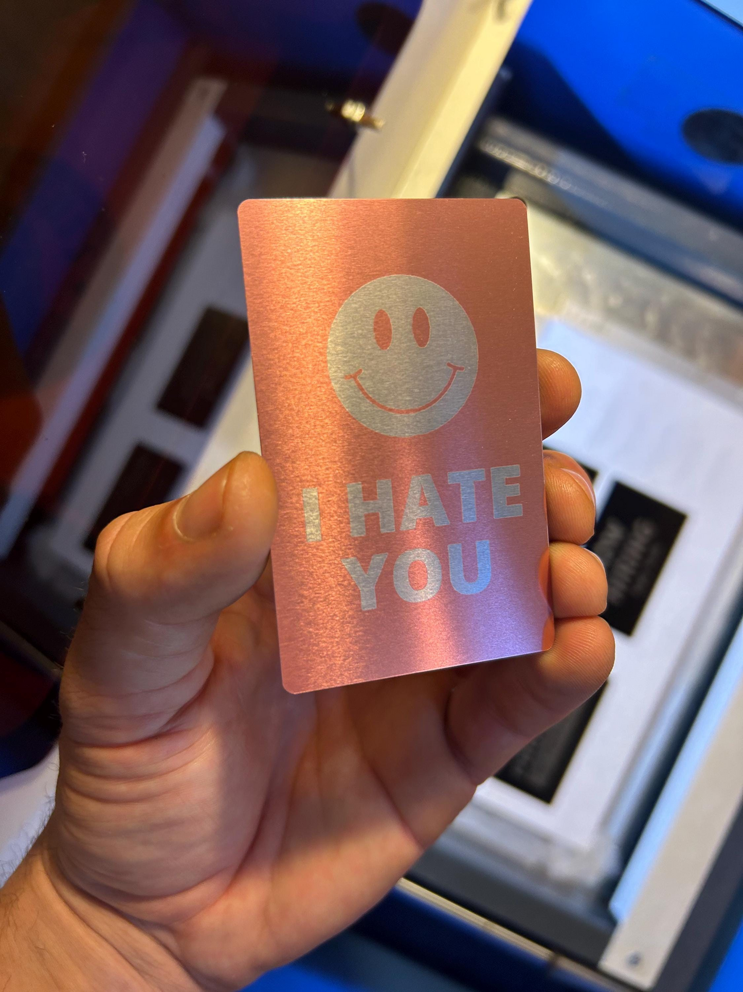 Hand holding an anodized aluminum gag gift card with a smiley face and 'I HATE YOU' text against a blurred indoor background.
