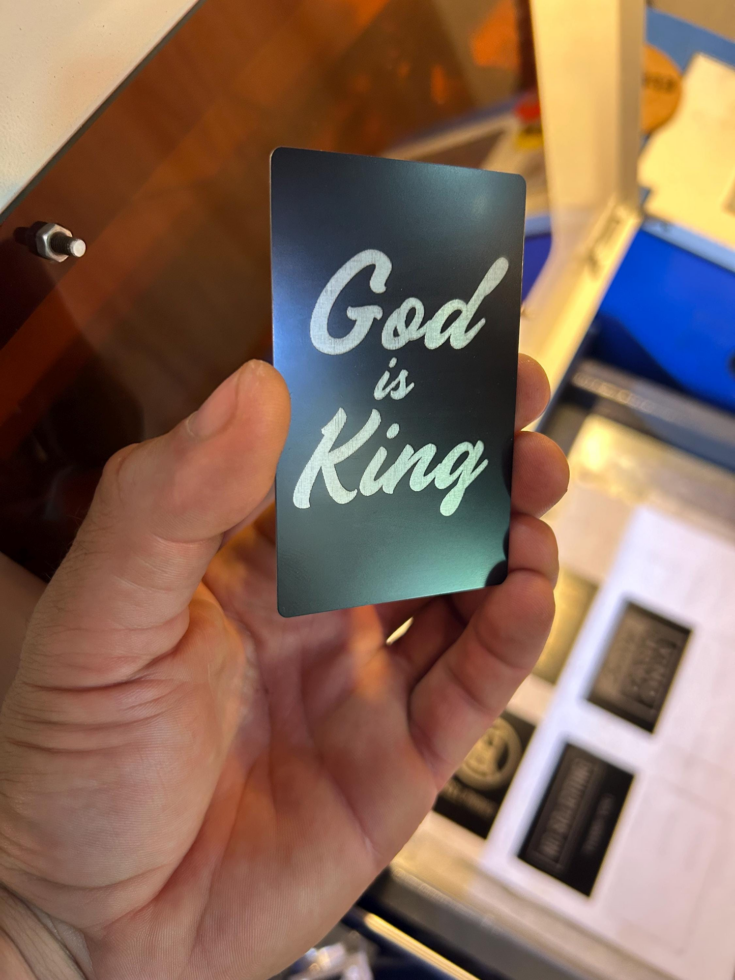Hand holding a metal black/silver card with 'God is King' engraved text against a blurred indoor background