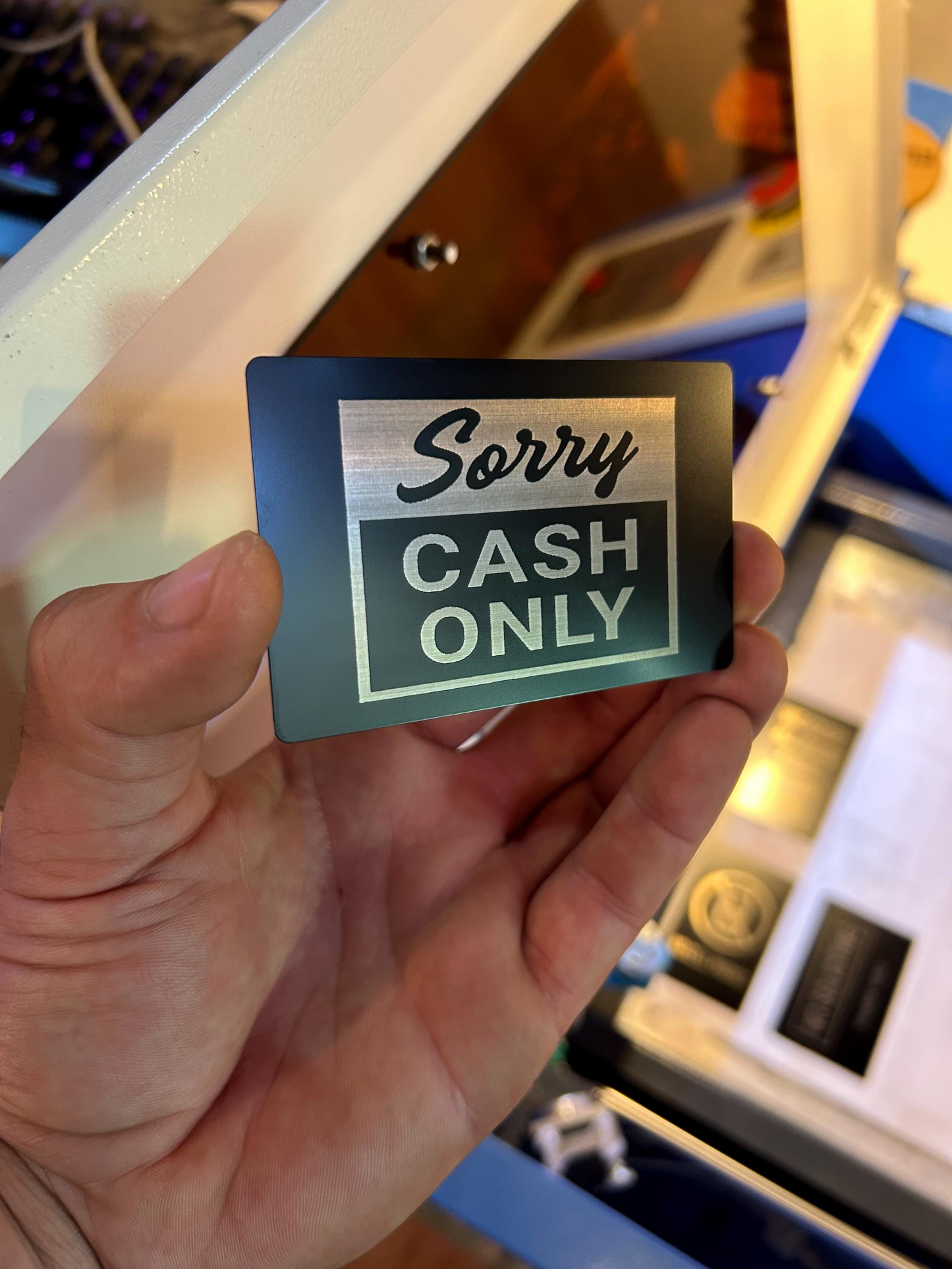 Hand holding a metal placard that says 'Sorry, Cash Only' in front of a blurred background.
