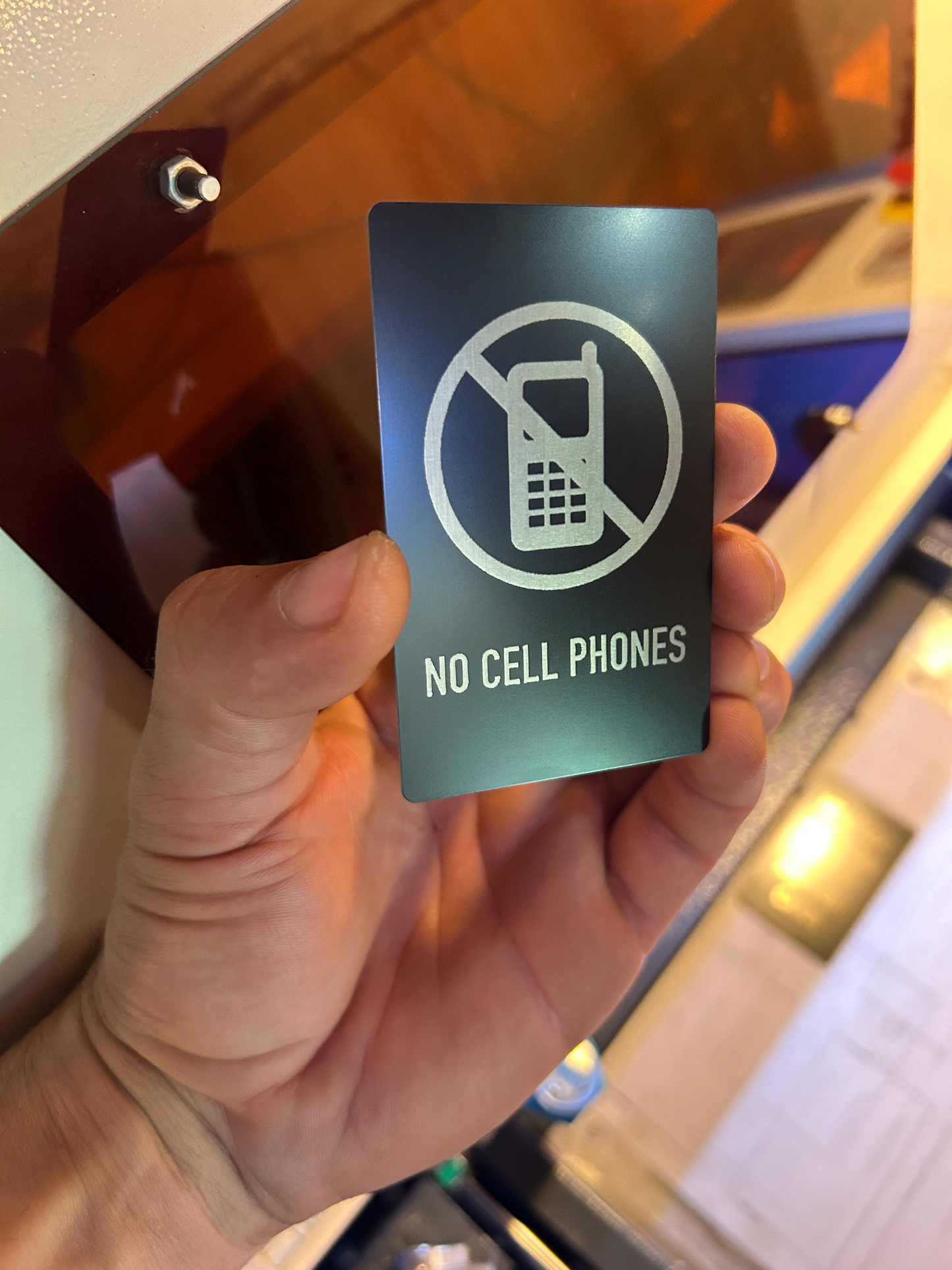 Hand holding a laser engraved anodized aluminum 'No Cell Phones' sign in black/silver in front of a machine