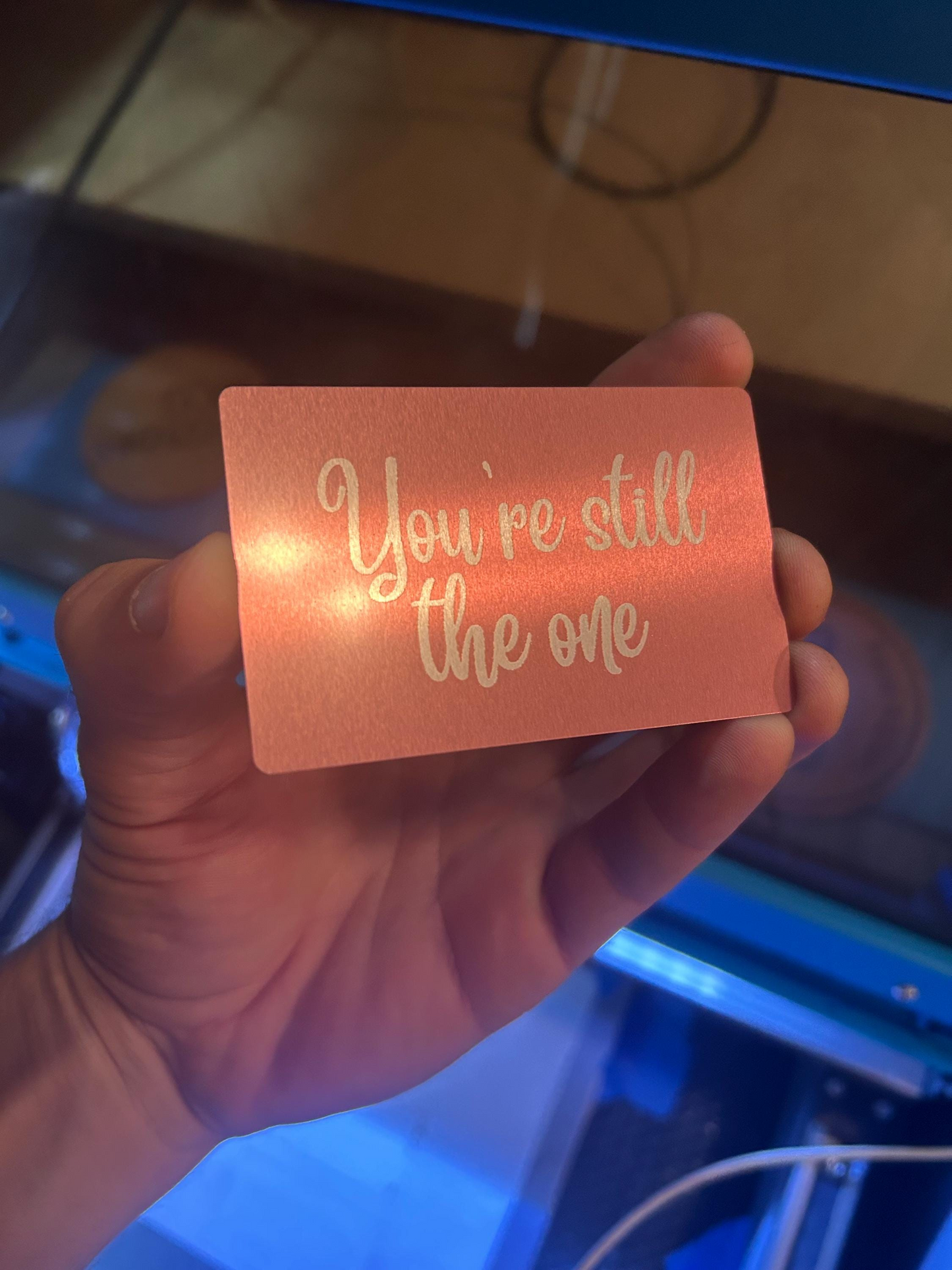Hand holding a rose gold/silver metal keepsake card with 'You're still the one' text in a softly lit indoor setting.
