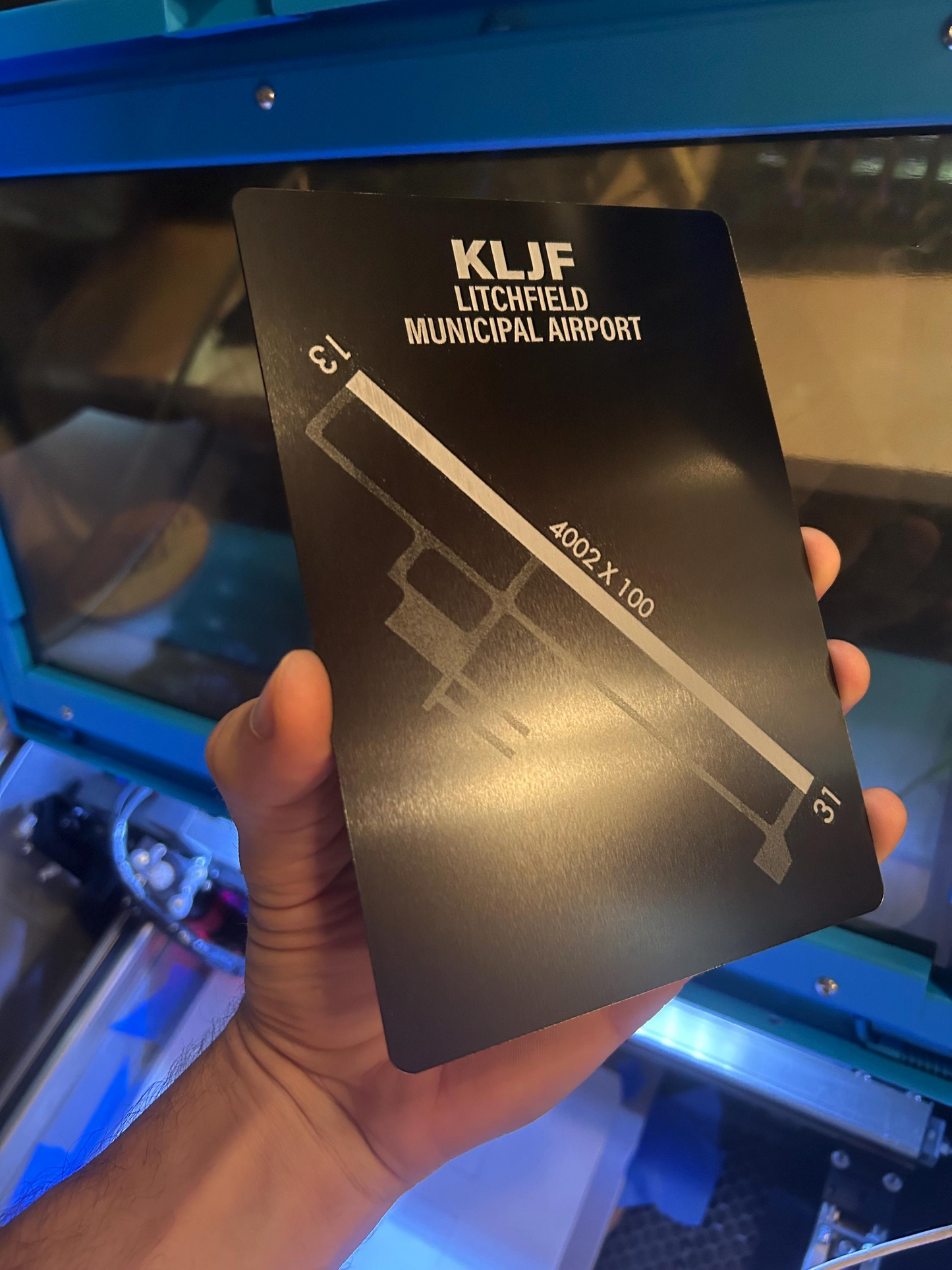 Decorative plaque with KLJF Litchfield Municipal Airport layout laser engraved in metal -- held by a hand in front of a blue machine.