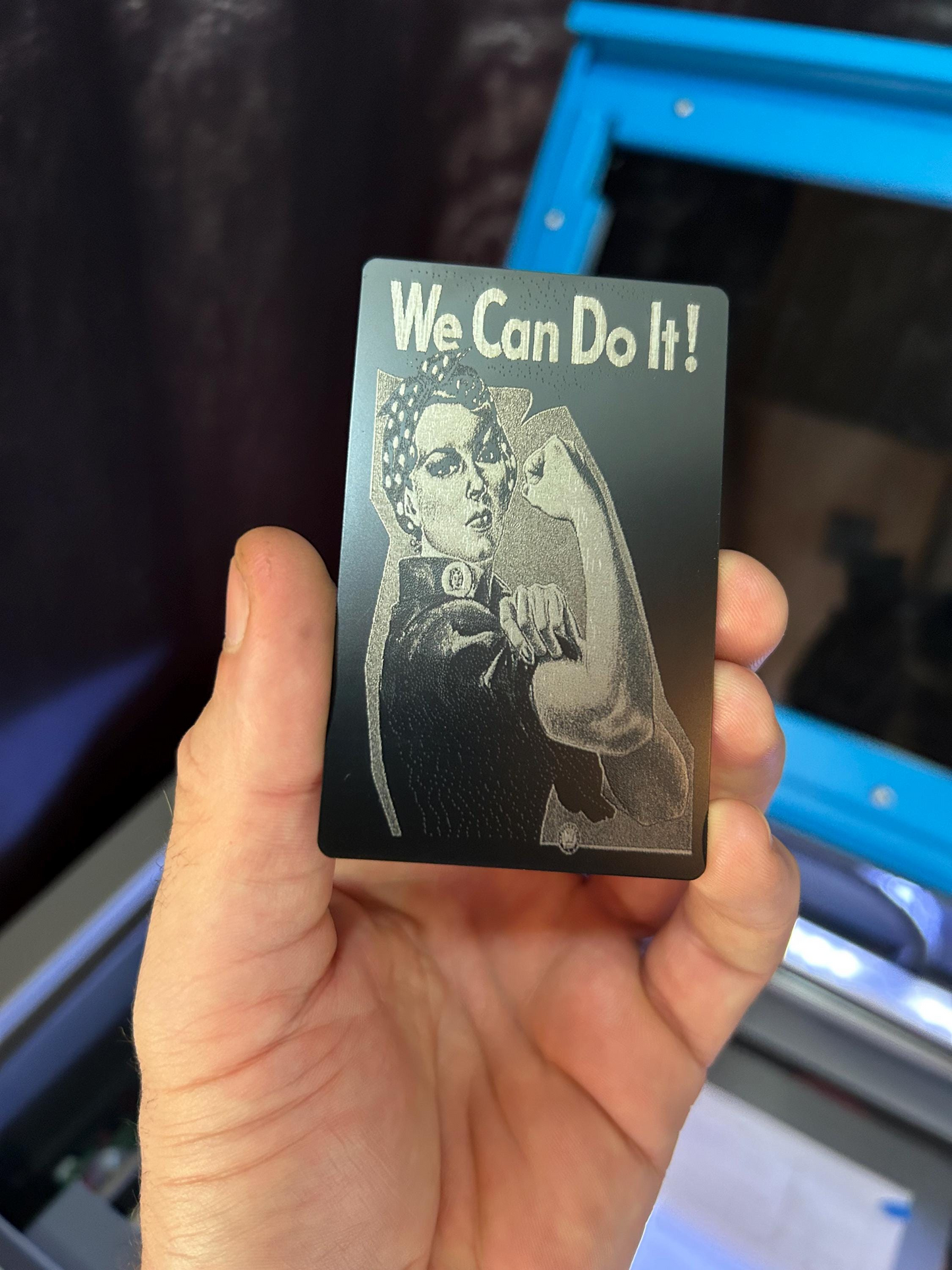 Hand holding a laser engraved metal gift card with 'We Can Do It!' text and a vintage-style laser engraved artwork