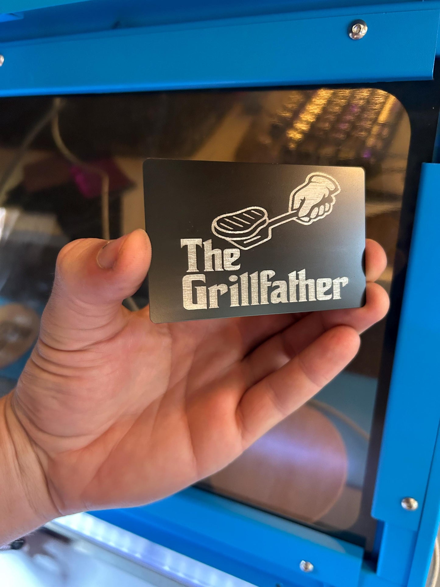 Hand holding a metal keepsake card with 'The Grill father' spatula burger design in front of a blue machine.