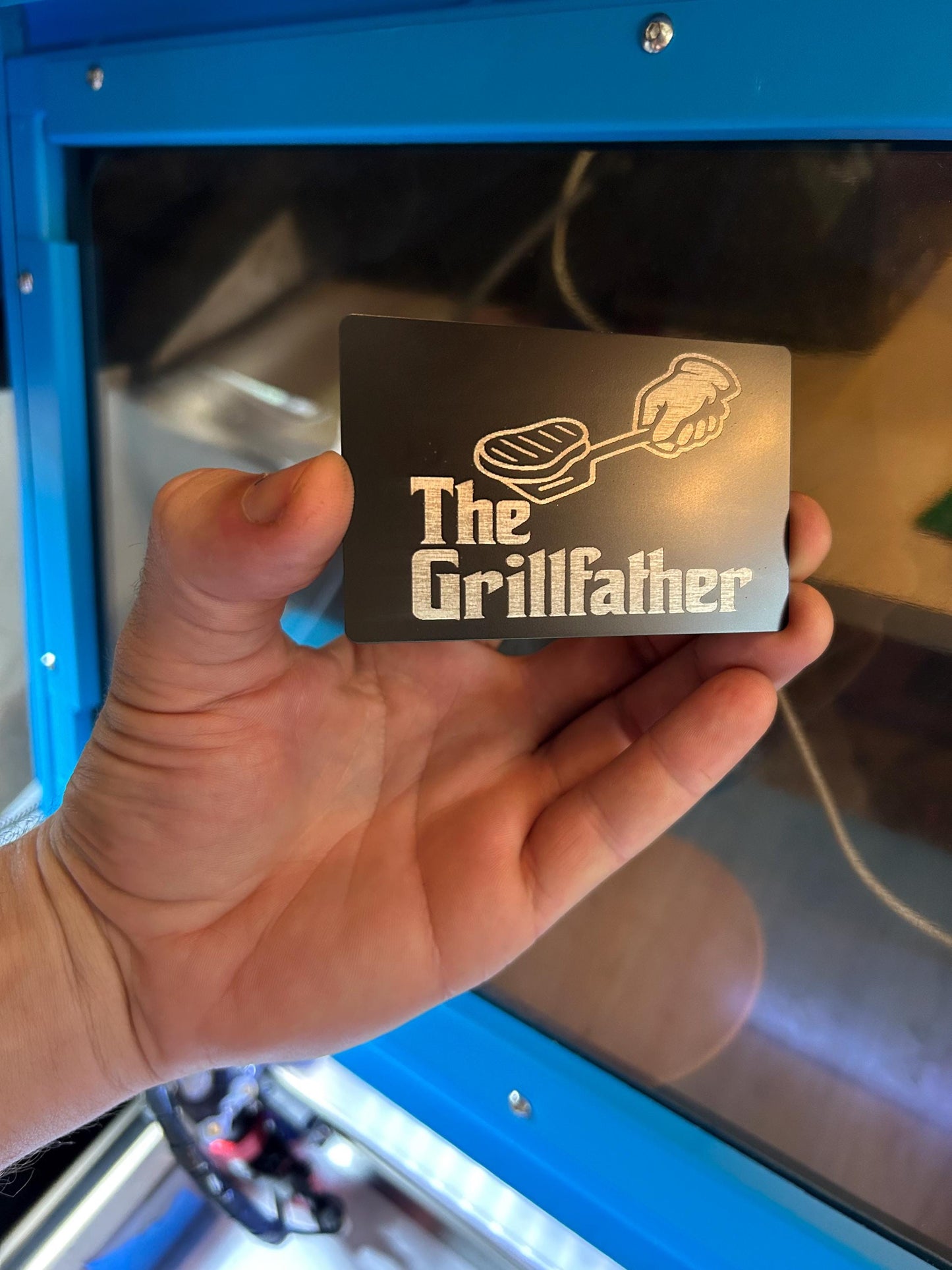 Hand holding a dad placard with 'The Grill father' design in front of a grill.