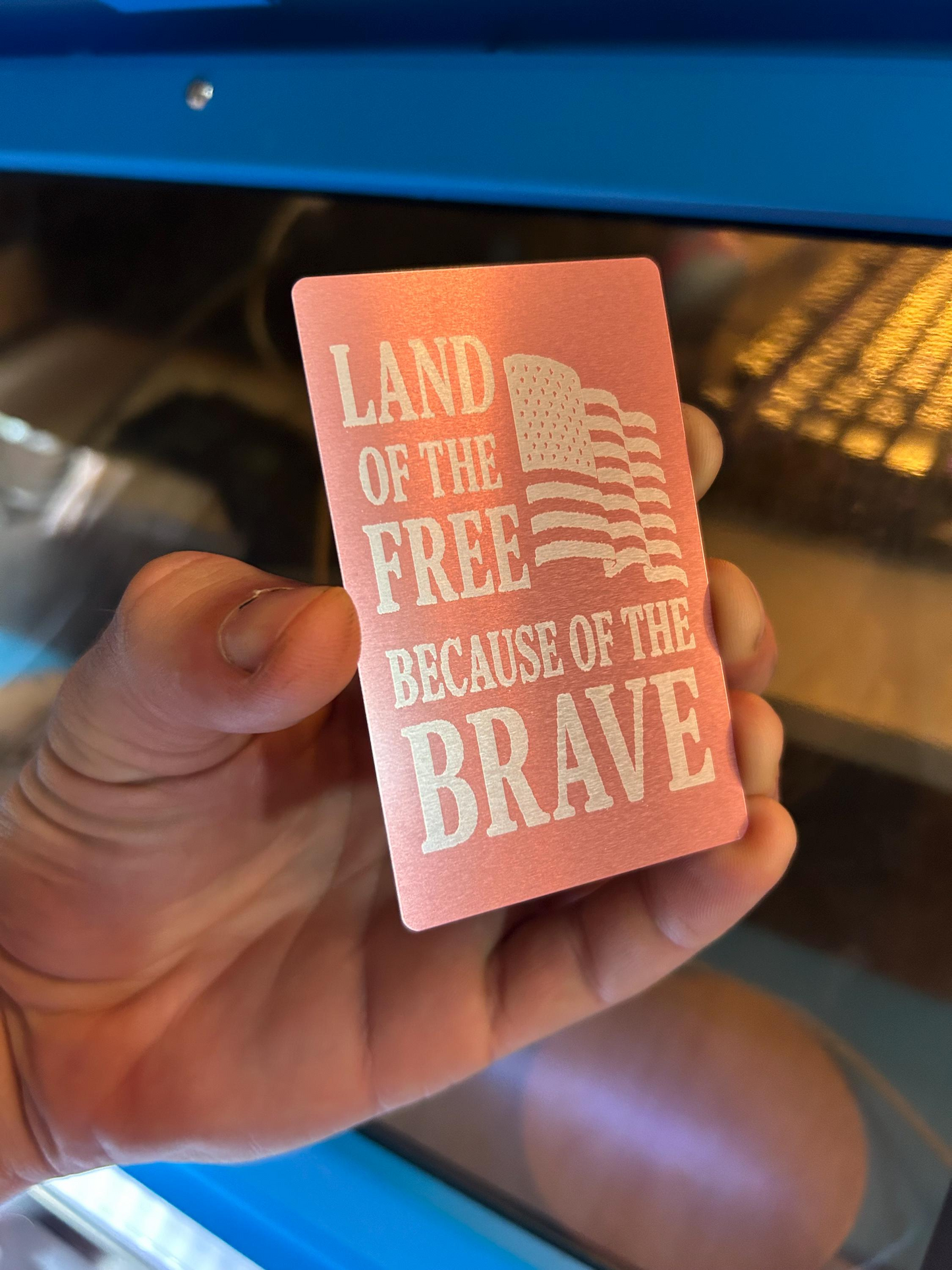 Hand holding a pink card with 'Land of the Free Because of the Brave' text and American flag design.