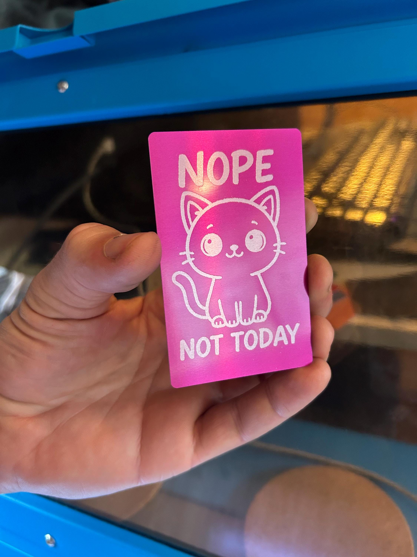 Hand holding a laser engraved placard with a cat illustration and text 'NOPE NOT TODAY' in front of a blue object.