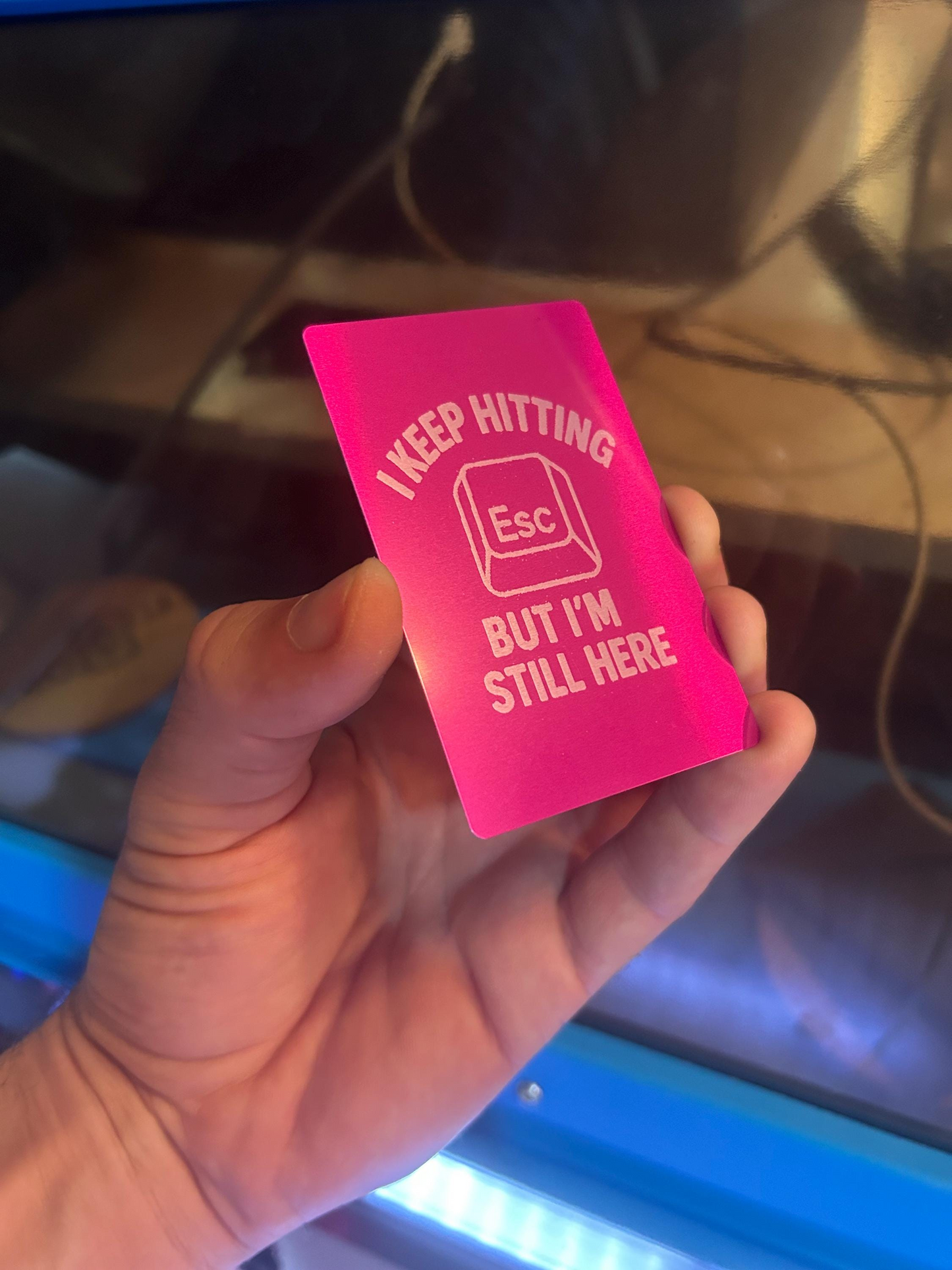 Hand holding a purple metal business card with personalized funny text and an icon, against a blurred laser cutting machine background.