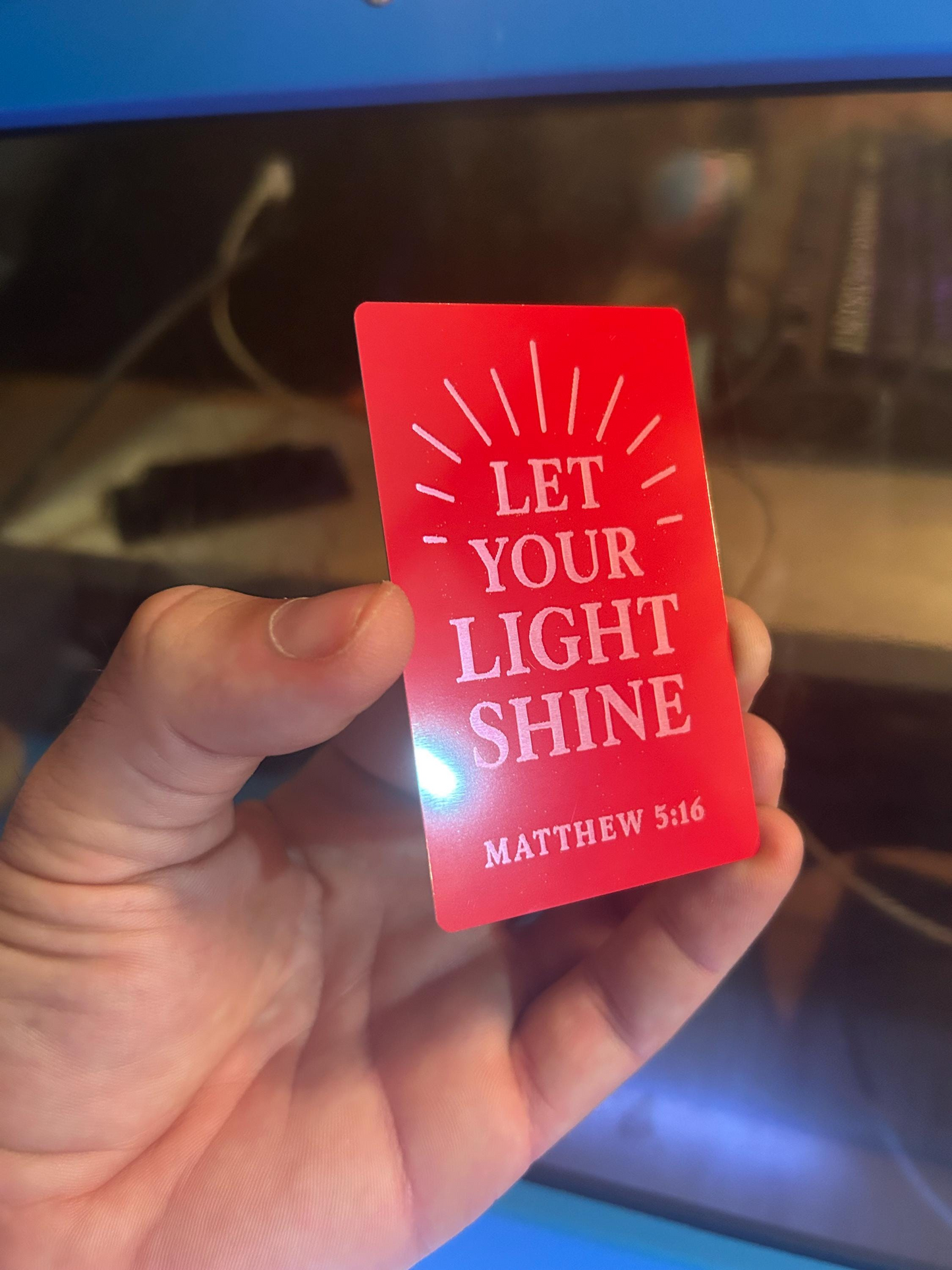 Hand holding a red and silver metal keepsake card with 'Let Your Light Shine' text with sun rays and Matthew 5:16 bible reference against a blurred laser machine background